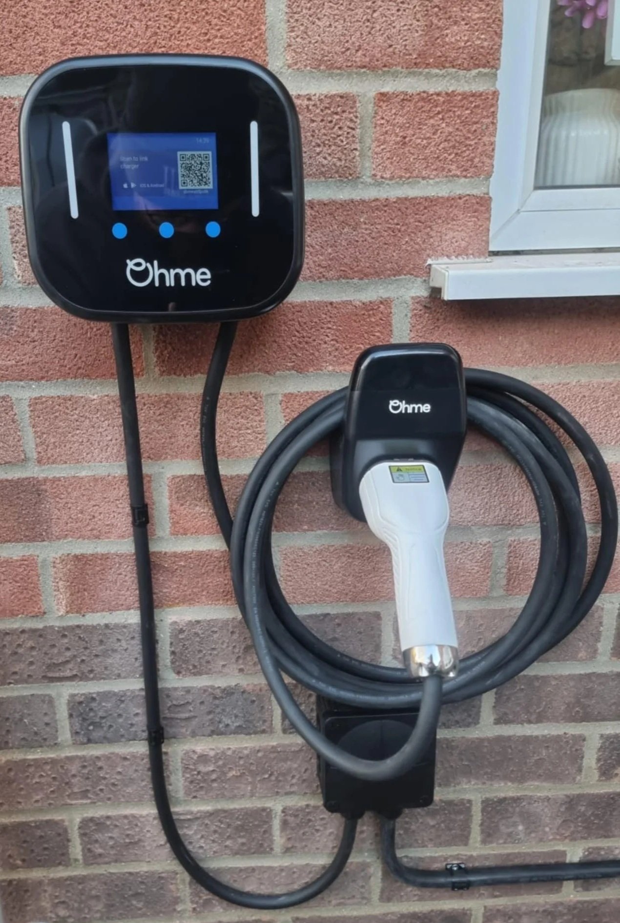 Electric vehicle charging station mounted on a brick wall, with a digital screen and a charging cable with a connector.