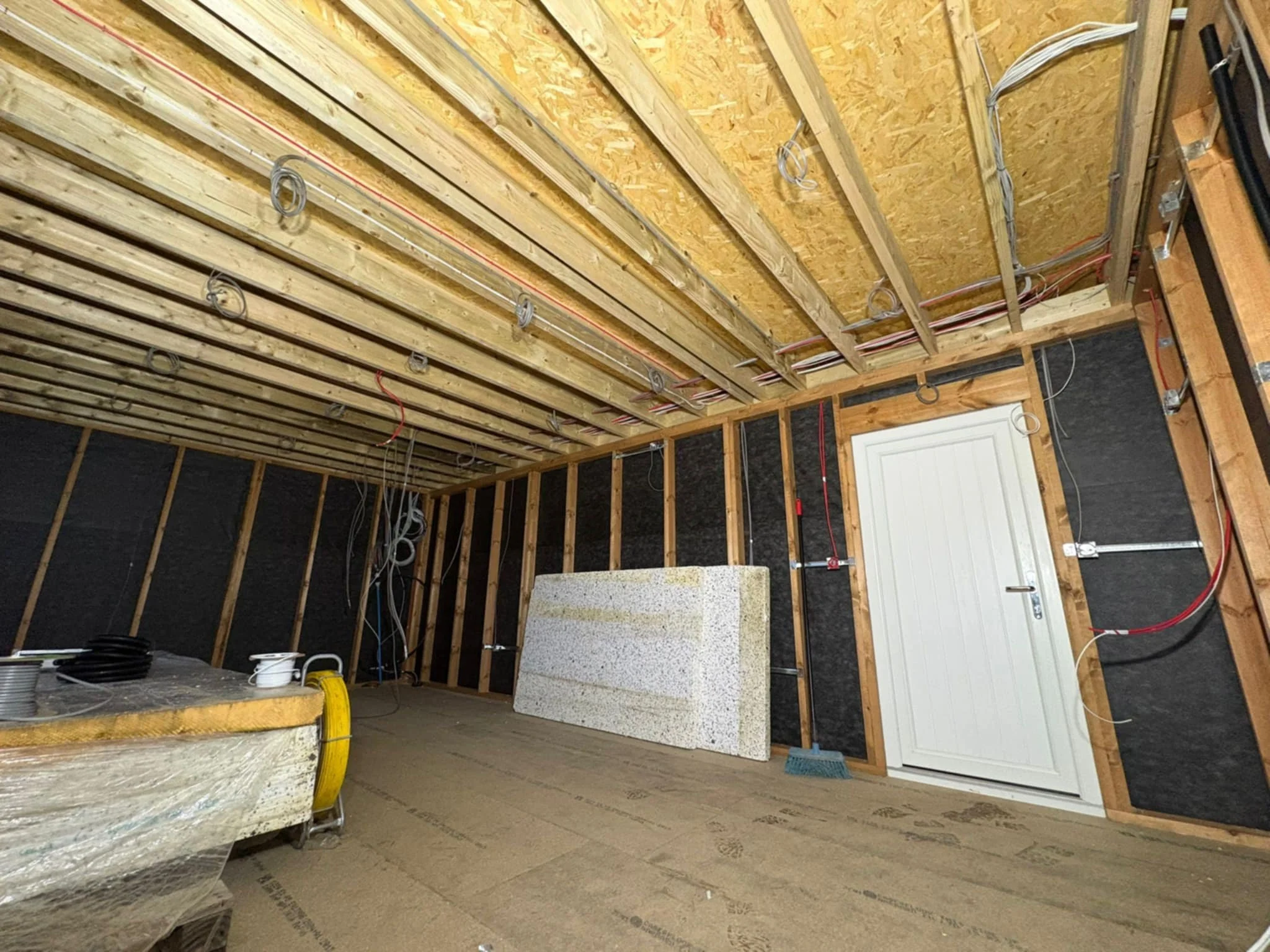 Construction site with exposed wooden framing, electrical wiring, and a door. A large foam block and construction materials are on the floor.