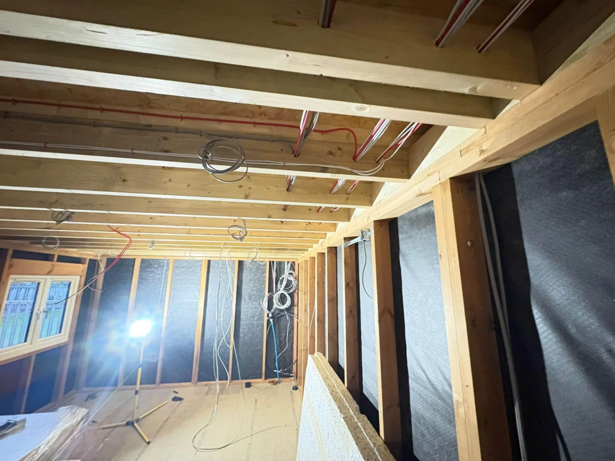 Interior of a room under construction, showing exposed wooden framing, electrical wiring, and some insulation on the walls. There is a window on the left side.