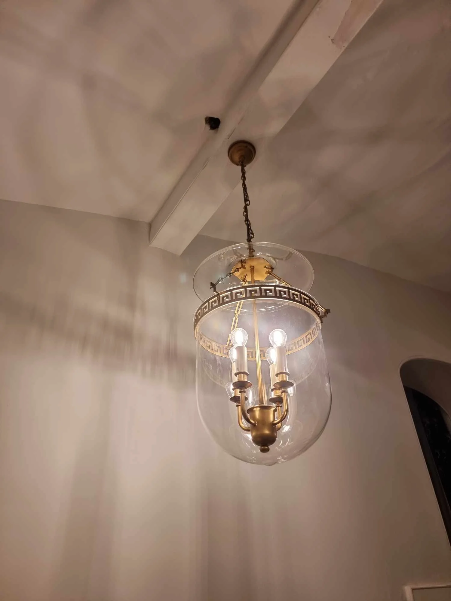 A chandelier with a brass finish and four light bulbs encased in a clear glass cover, hanging from a ceiling with visible beams.