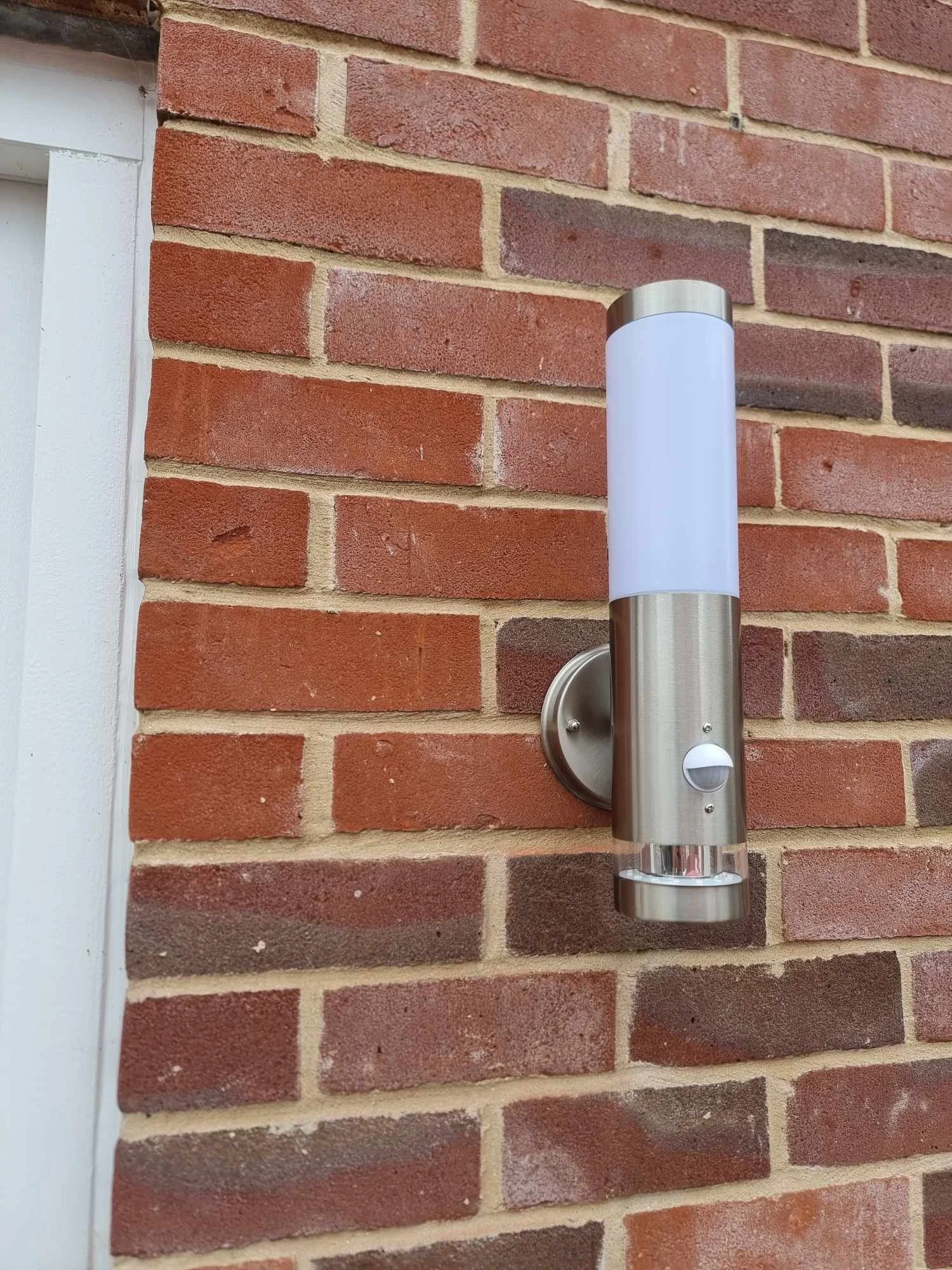 A modern outdoor wall-mounted light fixture with a cylindrical shape, stainless steel top and bottom, a frosted white middle section, and a small motion sensor at the bottom, mounted on a red brick wall.