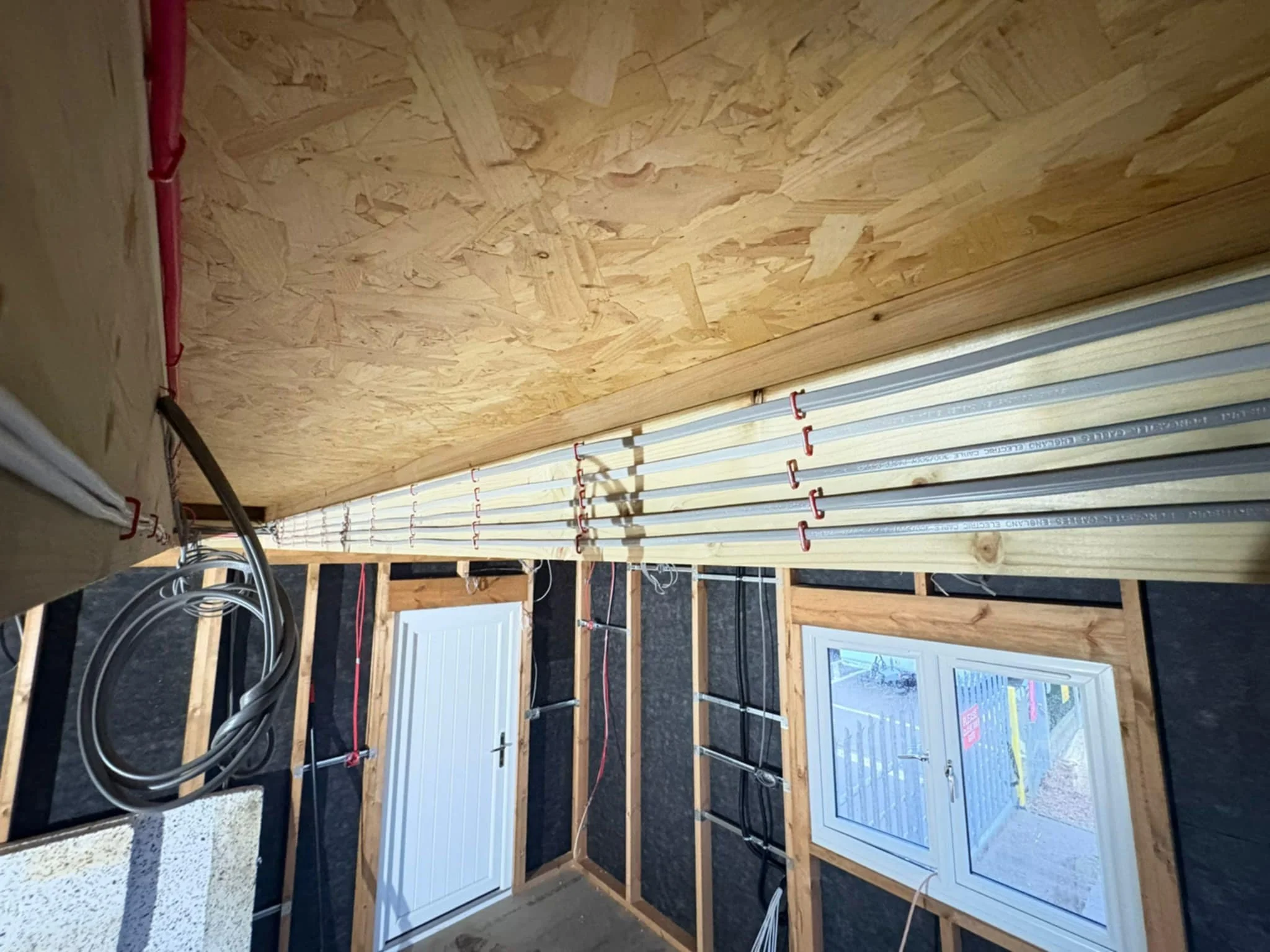 Construction site with wooden studs, pipes, and wiring, a window, and a door in an unfinished room.