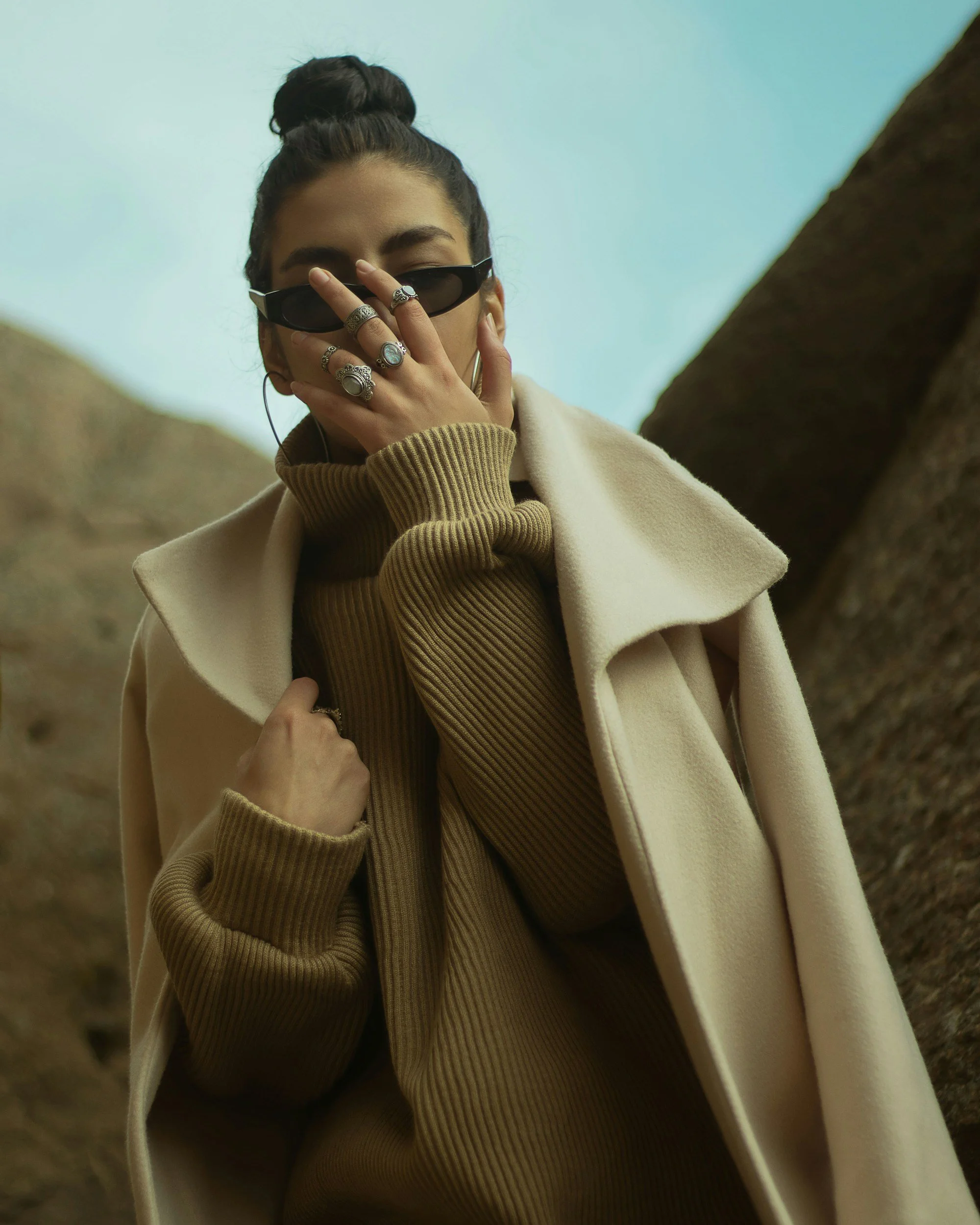 A woman with dark hair in a bun, wearing sunglasses, jewelry, a beige coat, and a brown ribbed turtleneck, standing outdoors near rocks.