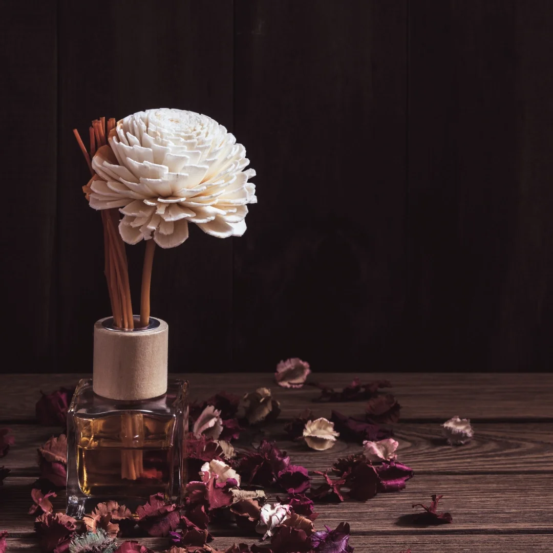 A soap flower diffuser with wooden sticks in a glass jar on a wooden surface, surrounded by dried flower petals.