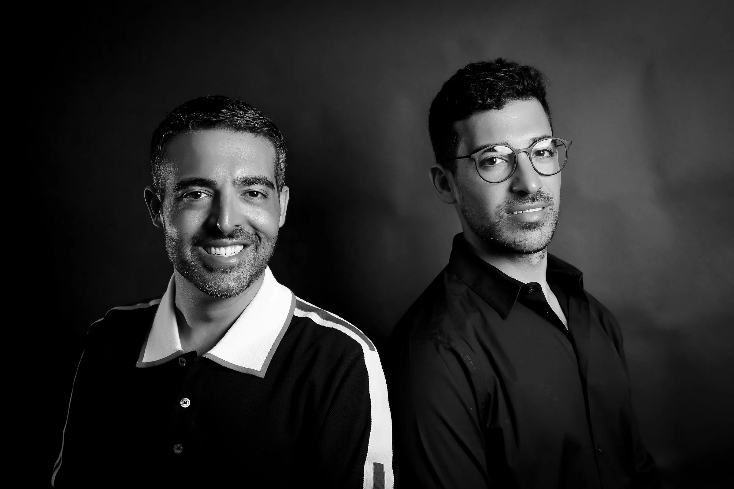 Black and white portrait of two men, one on the left smiling with short dark hair, wearing a polo shirt with white collar and stripes on shoulders; one on the right with glasses, short dark hair, wearing a collared shirt, both looking at the camera.