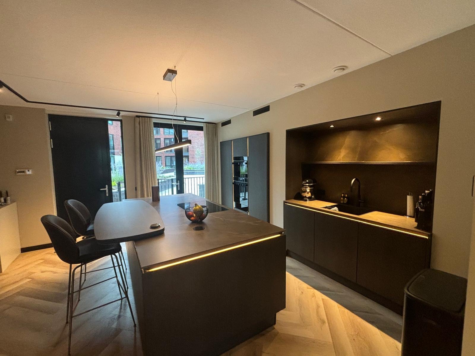 Moderne keuken en eetruimte met houten vloer, zwart keukenblok en grote keuken eettafel met drie zwarte barkrukken, uitzicht naar buiten door grote ramen met beige gordijnen.