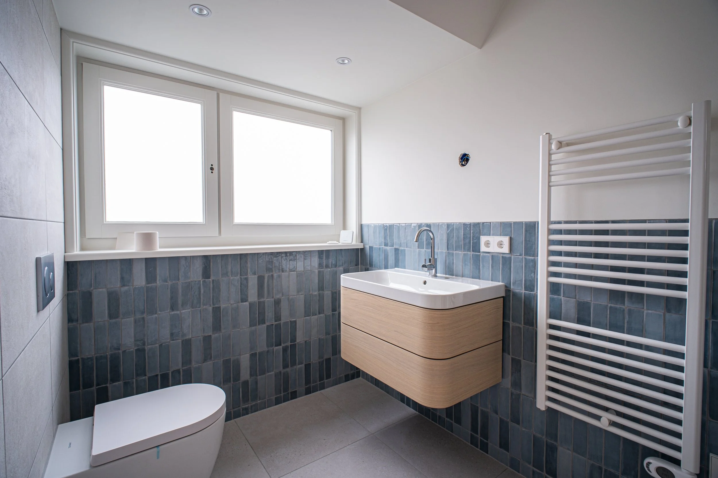 Een modern badkamer met een wit toilet, een zwart waterafvoersysteem, een witte wastafel met houten onderkast, een grote raam met wit kozijn en een witte handdoekradiator aan de muur.