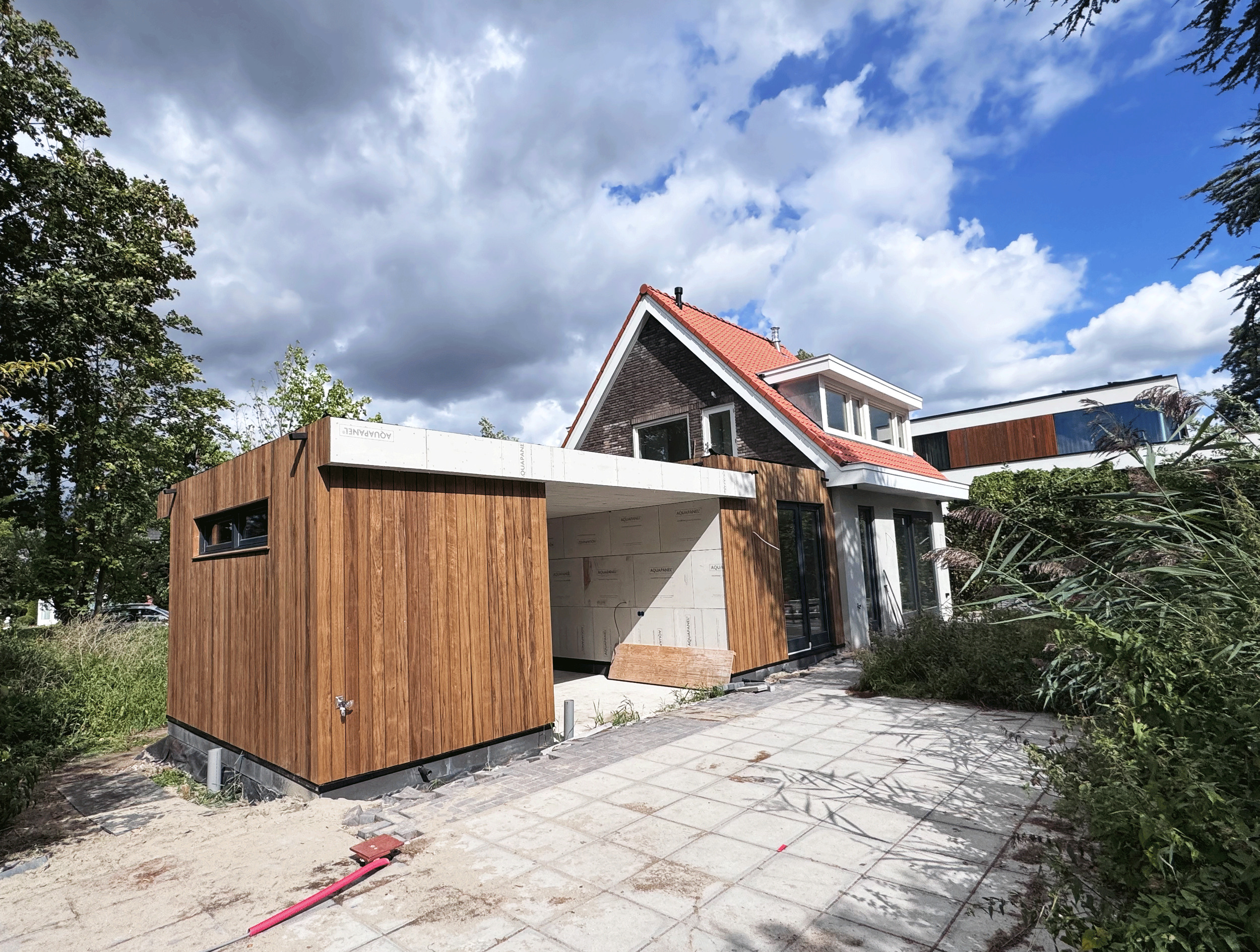 Nieuw modern huis onder constructie met houten en witte gevels, grote ramen, patio en tuin met planten, onder een bewolkte blauwe hemel