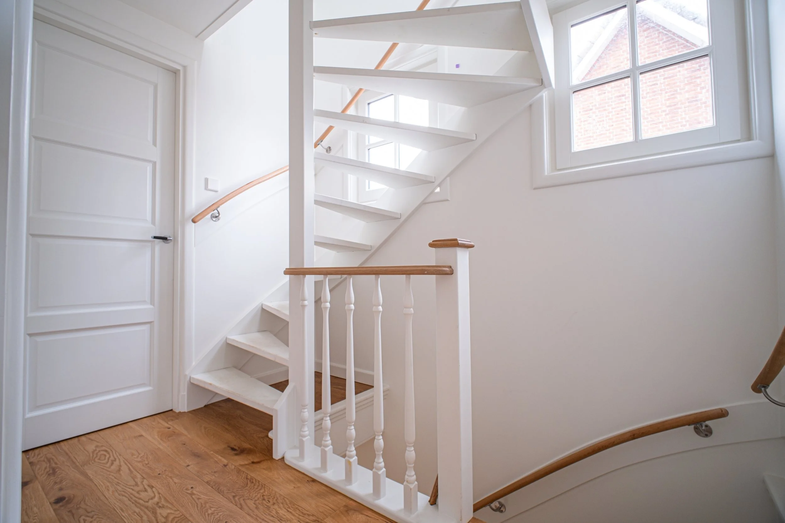Interieur van een huis met een witte wenteltrap en houten handrails, een open deur en twee ramen die natuurlijk licht binnenlaten.