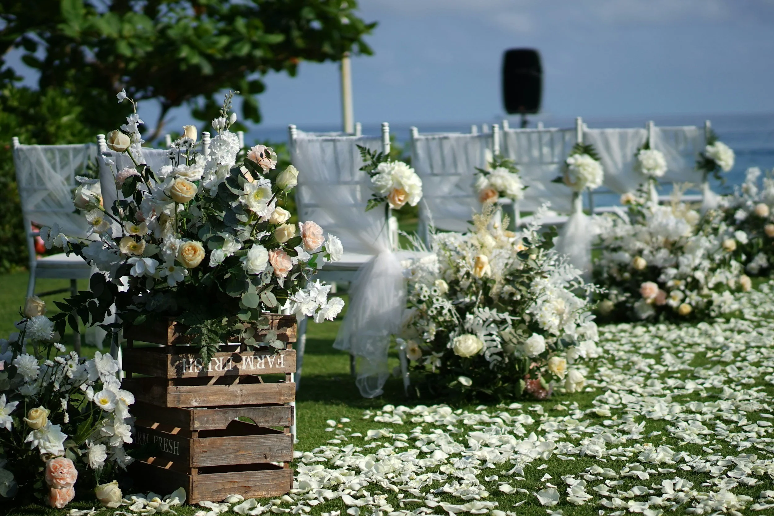 Outdoor wedding setup with white chairs, floral arrangements of white and pale pink flowers, white rose petals on green grass, near a body of water with blue sky, and a black speaker in the background.