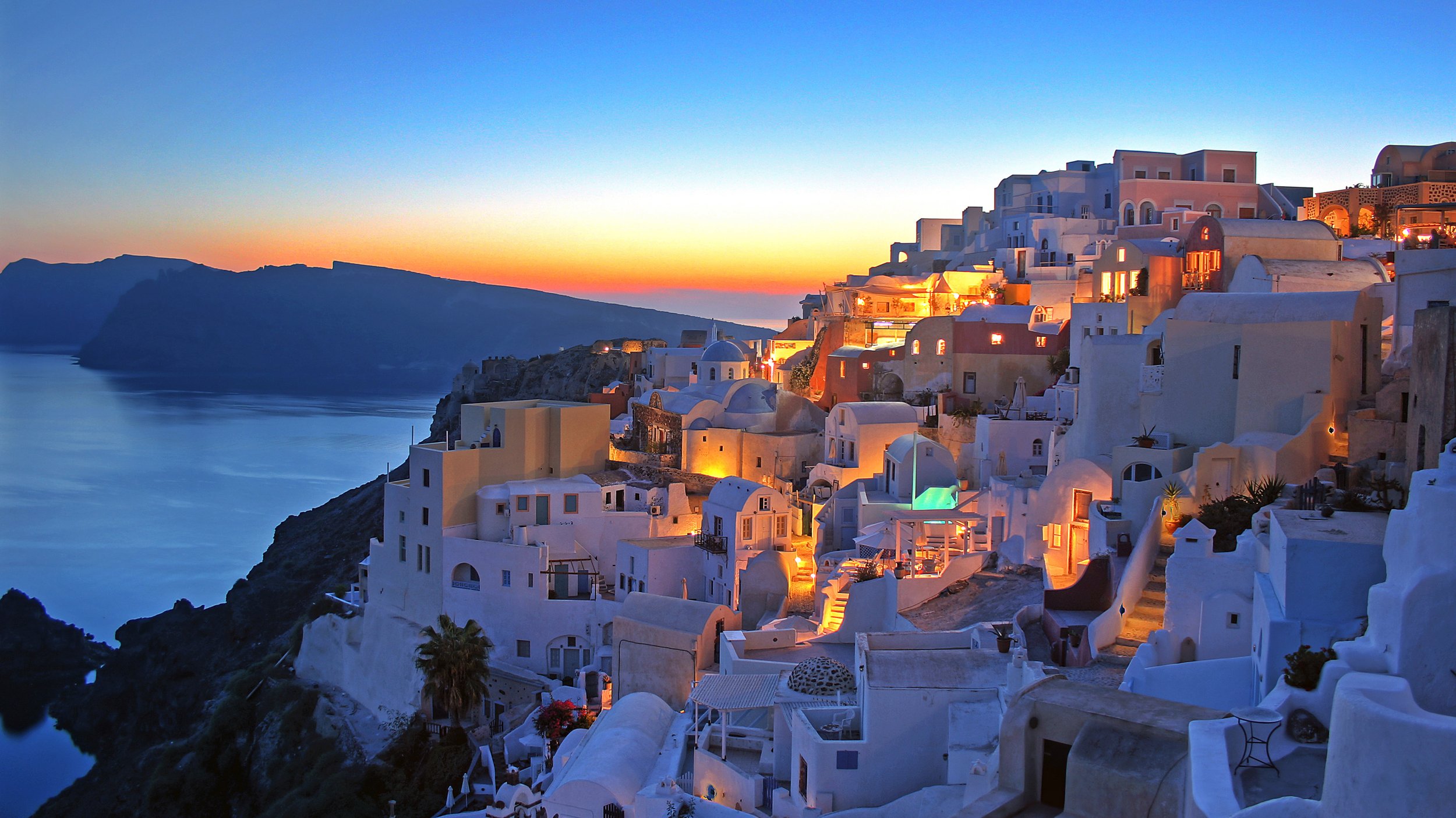 A picturesque view of a Greek island village at sunset with white buildings, narrow streets, and a calm sea in the background.