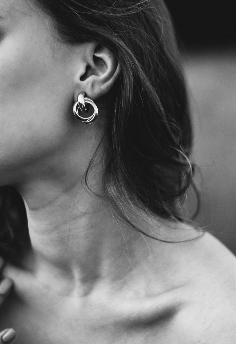 Close-up of a woman's left ear with a large, circular hoop earring, her shoulder and part of her neck visible, with wavy hair partially covering her ear in black and white.