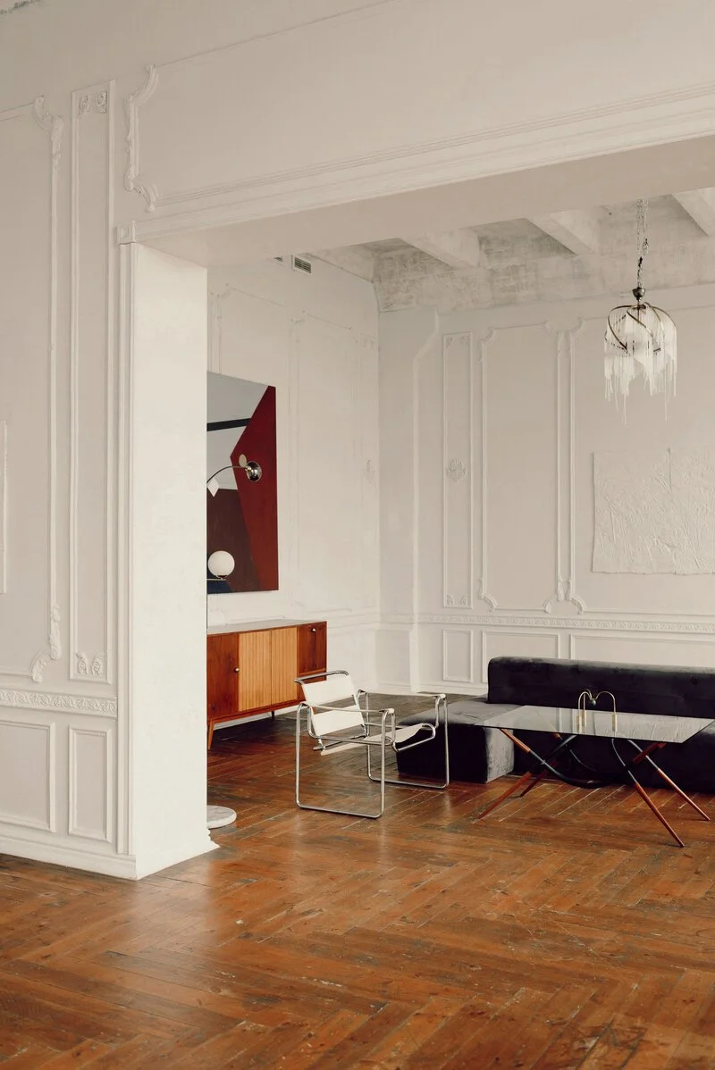 Minimalist living room with white ornate walls, wood flooring, a black sofa, a marble-top coffee table with wooden legs, two white modern chairs, a vintage wooden cabinet, an abstract wall art piece, and a chandelier with hanging glass pieces.