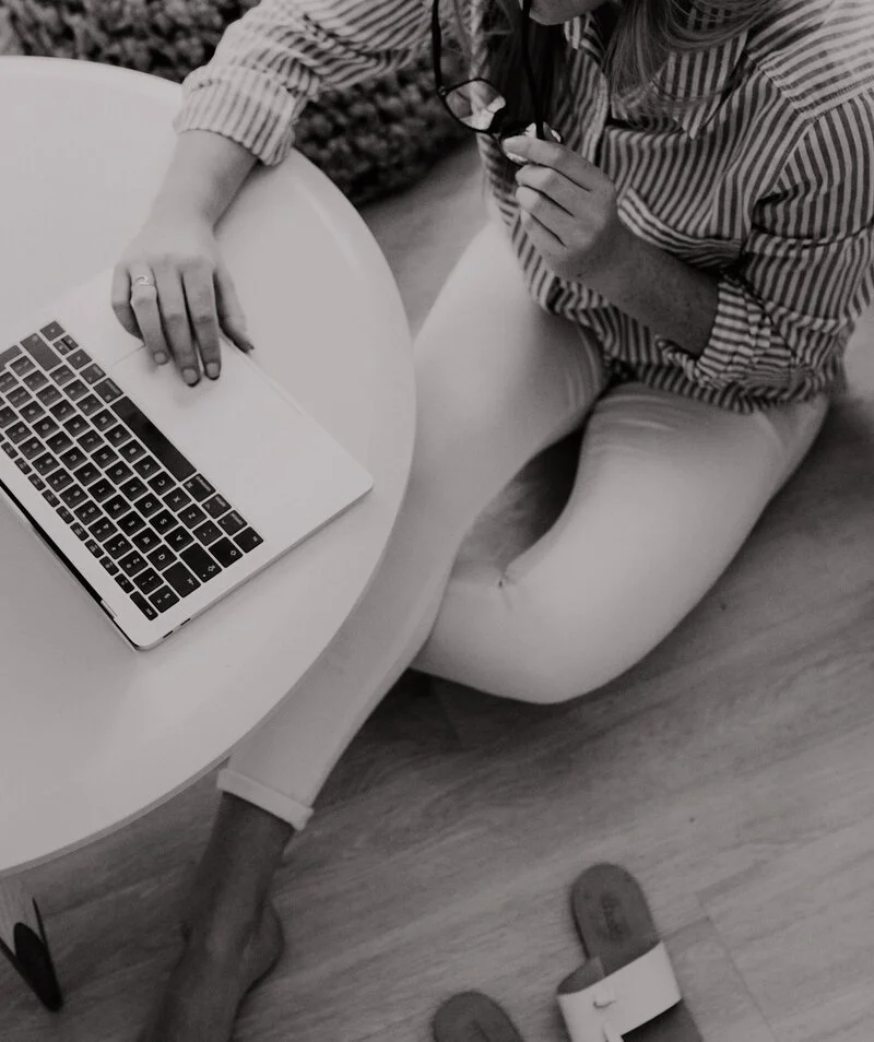A person sitting on the floor at a small round table with a laptop and glasses, wearing a striped shirt and light-colored pants, with sandals on the floor.