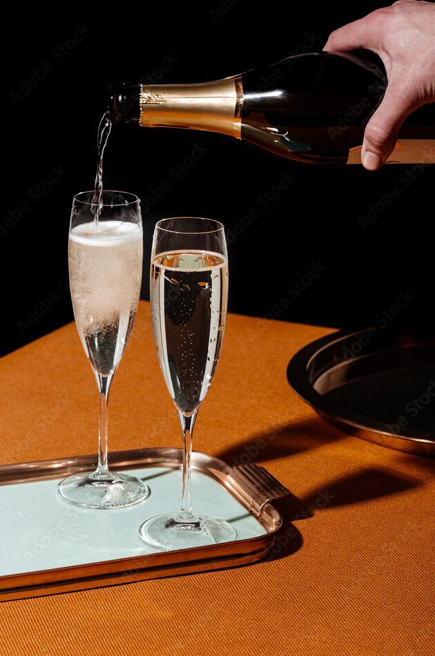 A hand pouring champagne into a flute glass on a table with a tray and a black background.
