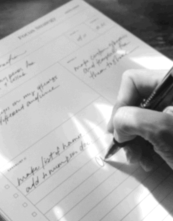 Close-up of a person's hand writing in a notebook with a pen, appearing to make a checklist.