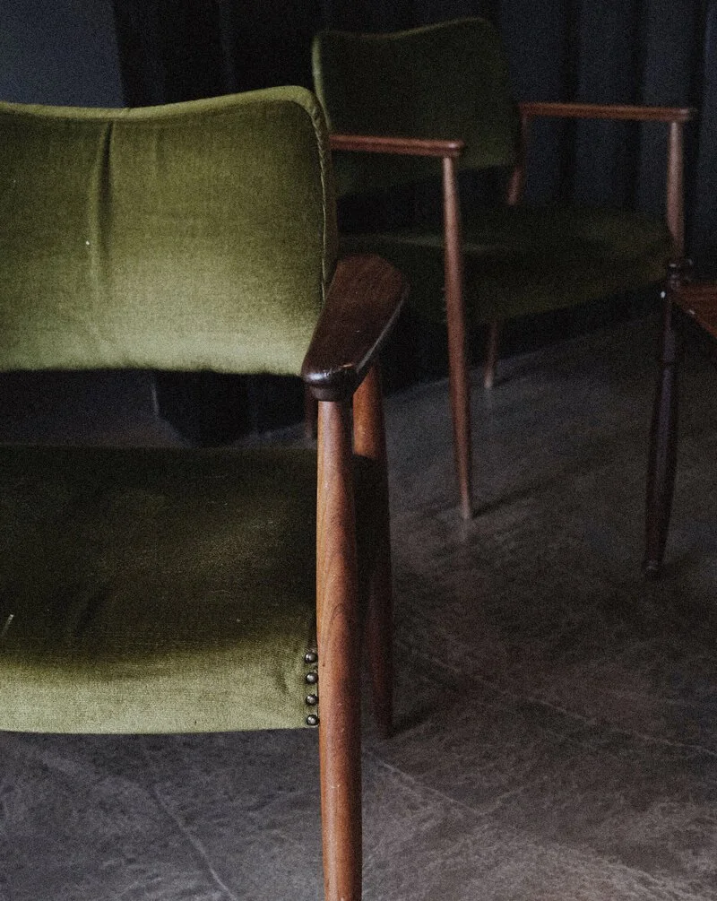 Two vintage green upholstered chairs with wooden armrests and legs, placed on a dark tiled floor.