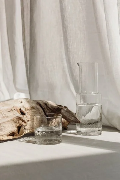 A glass of water, a tall beaker with water, and a piece of driftwood on a white surface in front of a white curtain.
