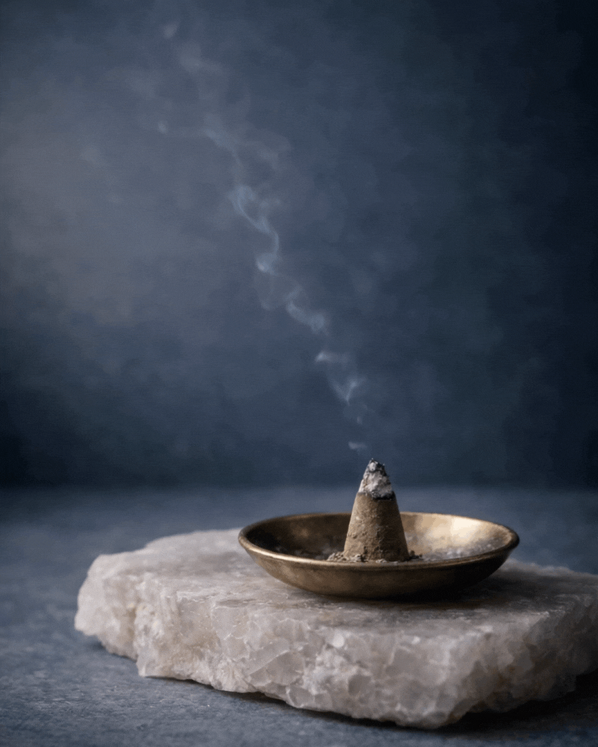 A smoldering cone incense placed in a small metal dish on a rough stone surface with smoke rising from it against a dark background.