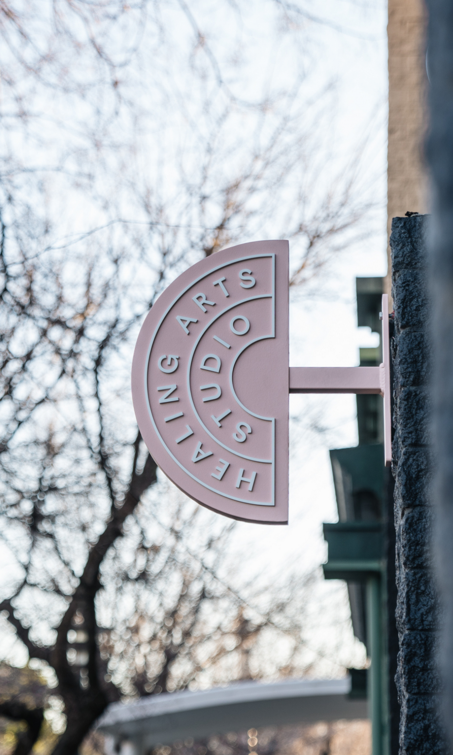 Pink circular sign with cut-outs of text that reads "HEALTH & ARTS AUDIO STUDIOS," attached to a stone wall outside. Blurred trees and a building are visible in the background.