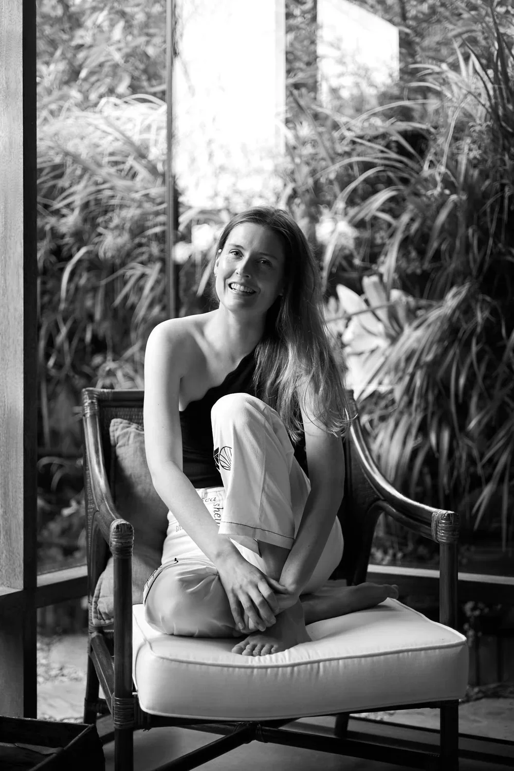 A woman sitting on a cushioned chair in a room with large windows, surrounded by tropical plants outside, smiling at the camera.
