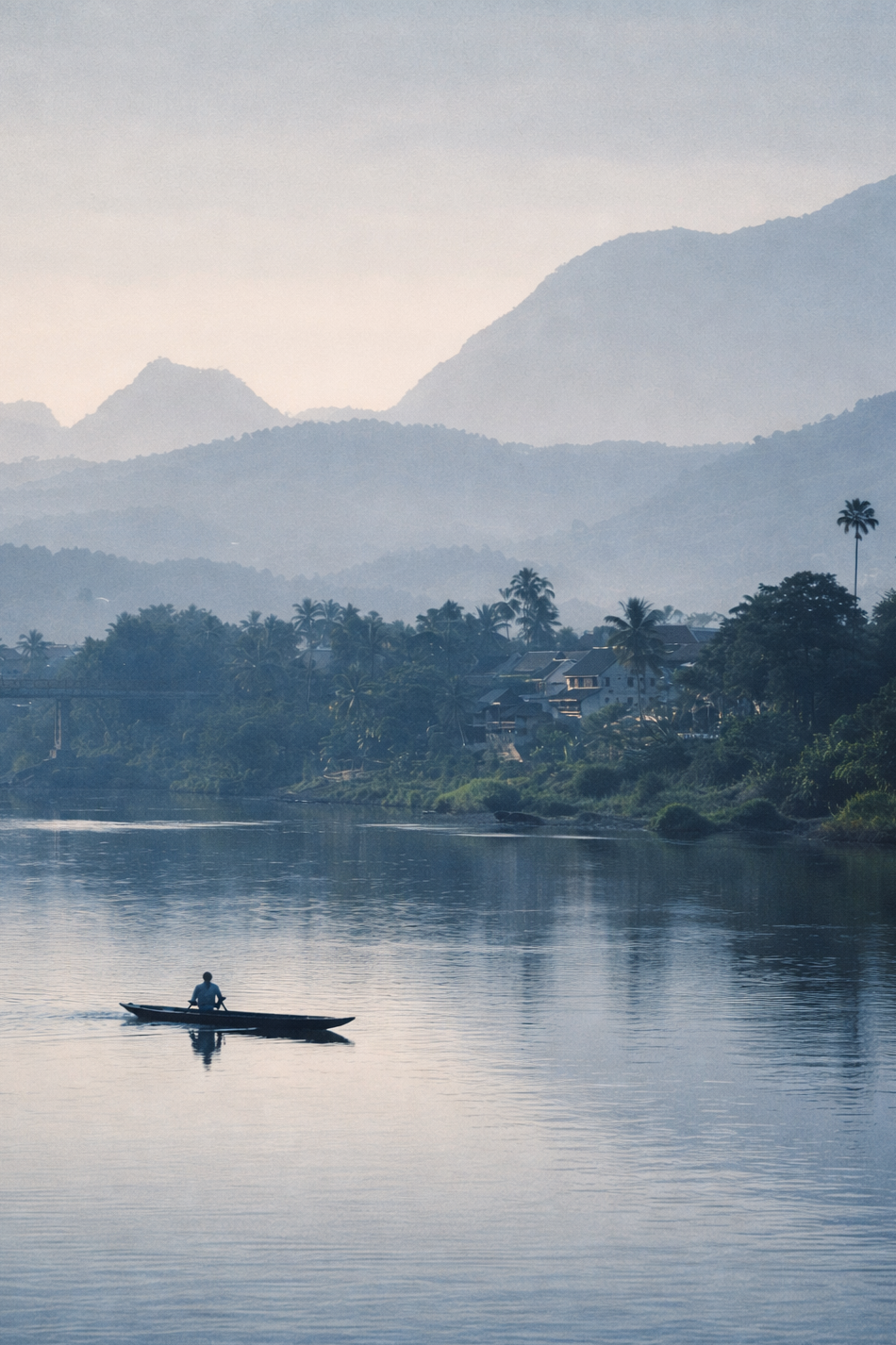 A person paddling a small boat on a calm river with lush greenery and houses along the shoreline, mountains in the background with a hazy sky.