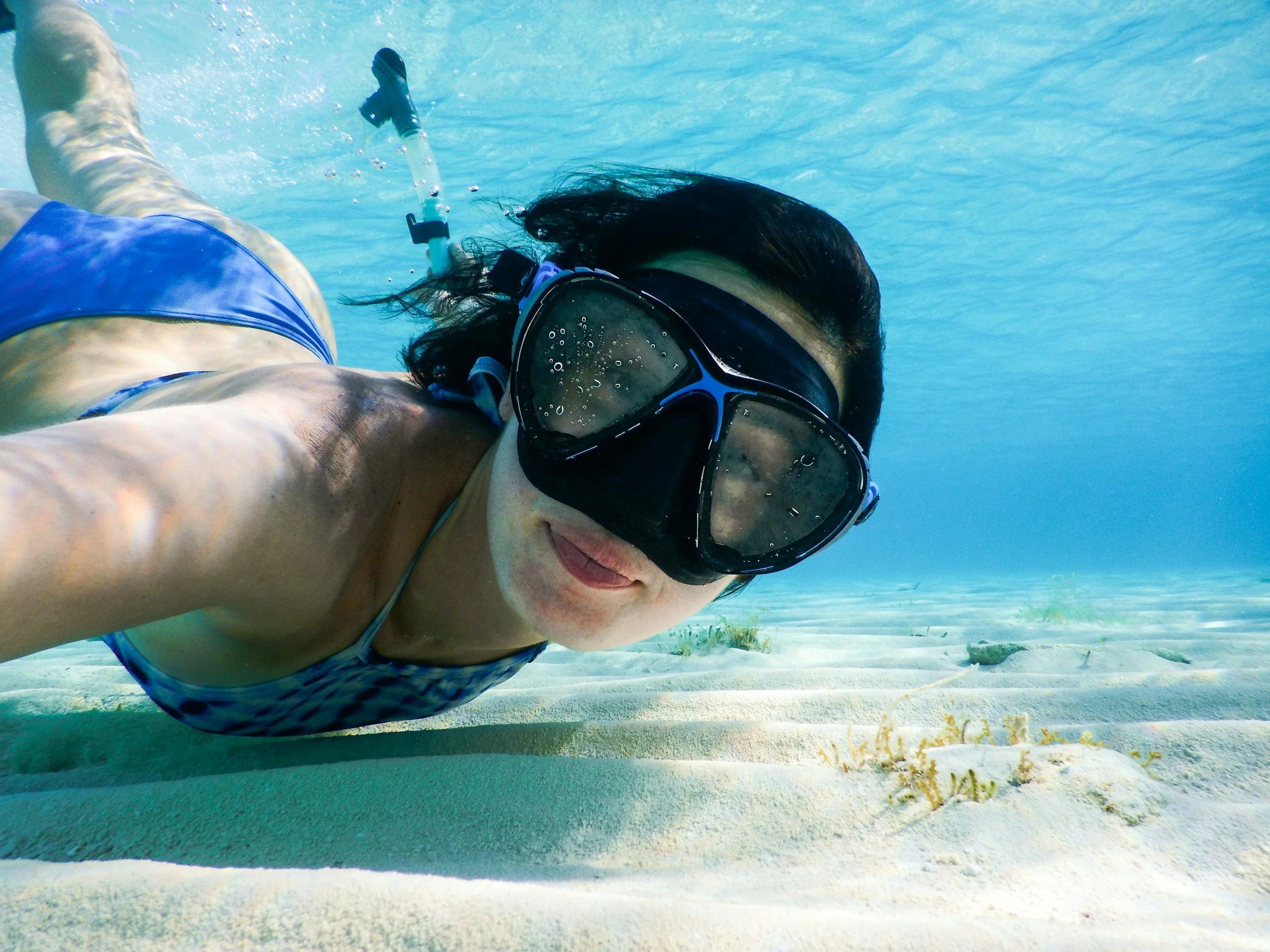 Woman swimming underwater in a swimsuit and snorkeling gear, close-up in clear blue water with sandy ocean floor and some sea plants.