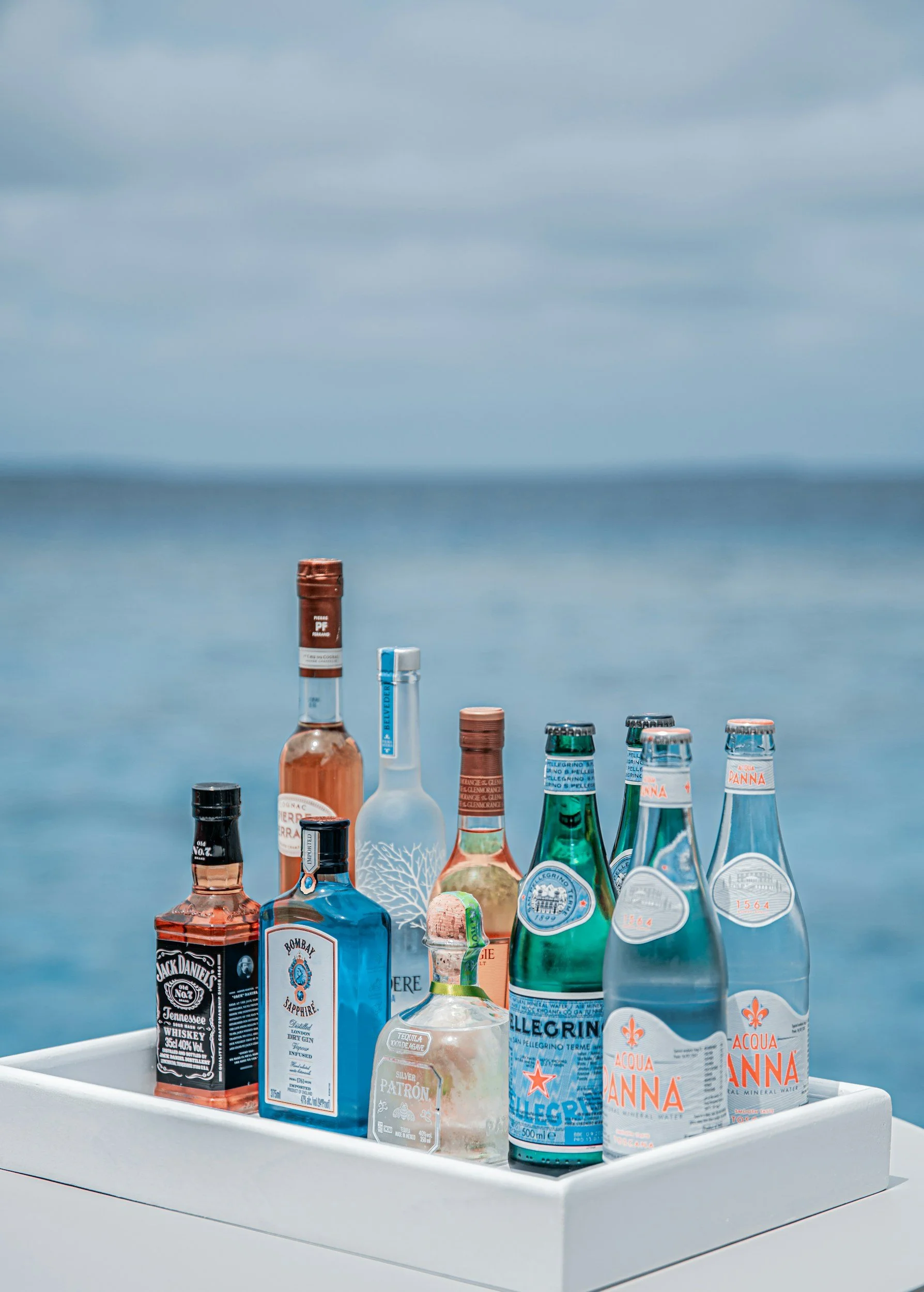 Assorted bottles of alcohol and water on a white tray against a blurred ocean background.