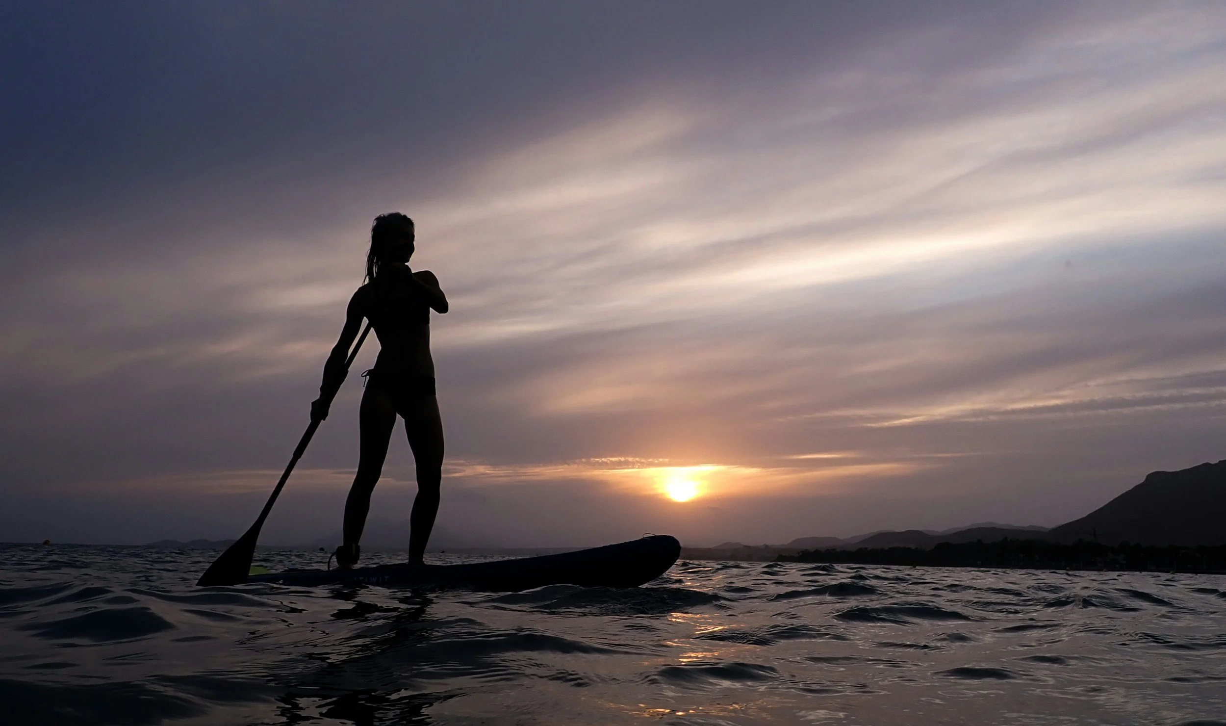Silhouette of a woman paddleboarding on the water at sunset, with mountains in the background and a cloudy sky.