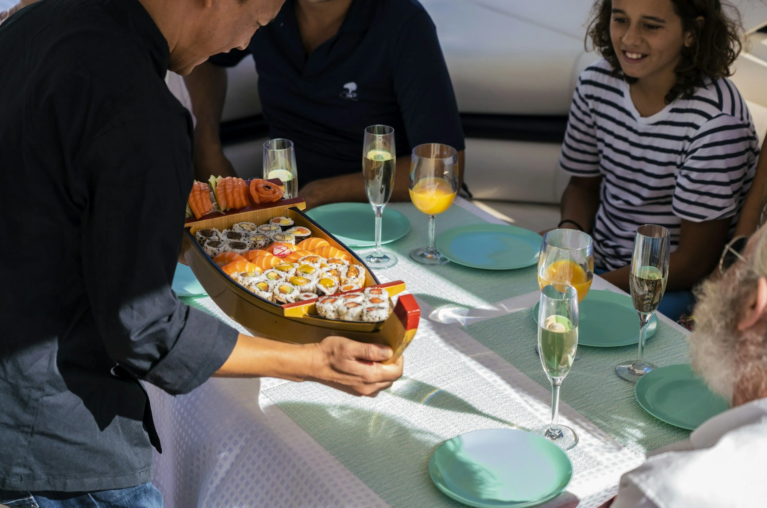 A man holding a boat-shaped tray of sushi while sitting at a decorated outdoor dining table with champagne glasses and turquoise plates during daytime.
