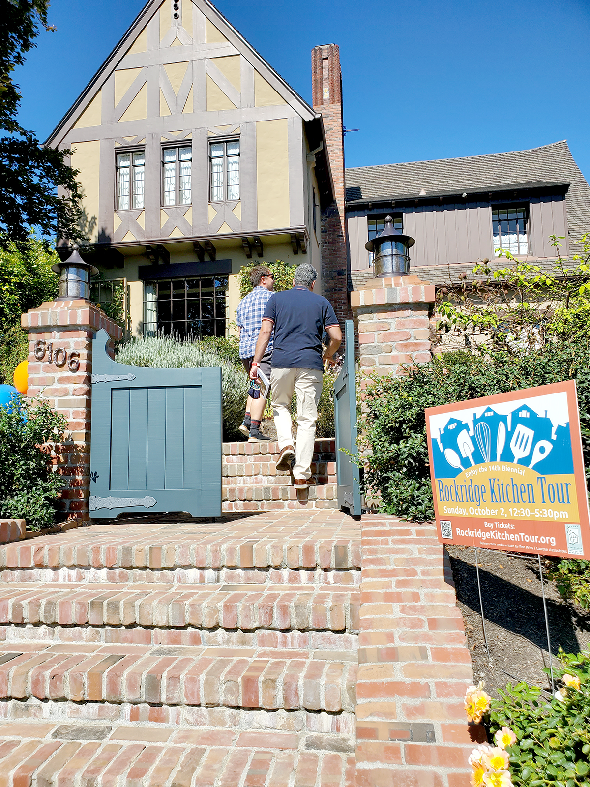 People entering a house for a kitchen tour event, with a sign in front indicating the date and tour details.