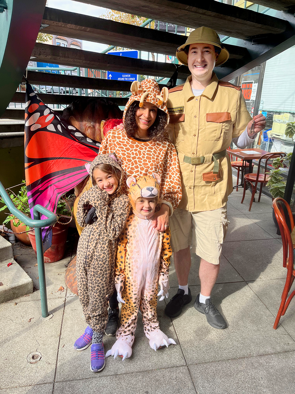 Family dressed in safari and animal costumes, including a giraffe, leopard, and lion, standing outdoors under a staircase.