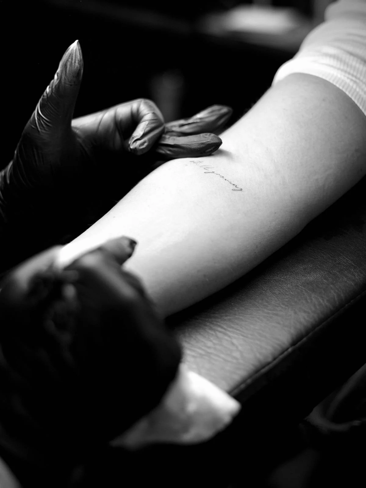 A person with gloves is tattooing a small script text on a person's arm, which is resting on a cushioned surface.