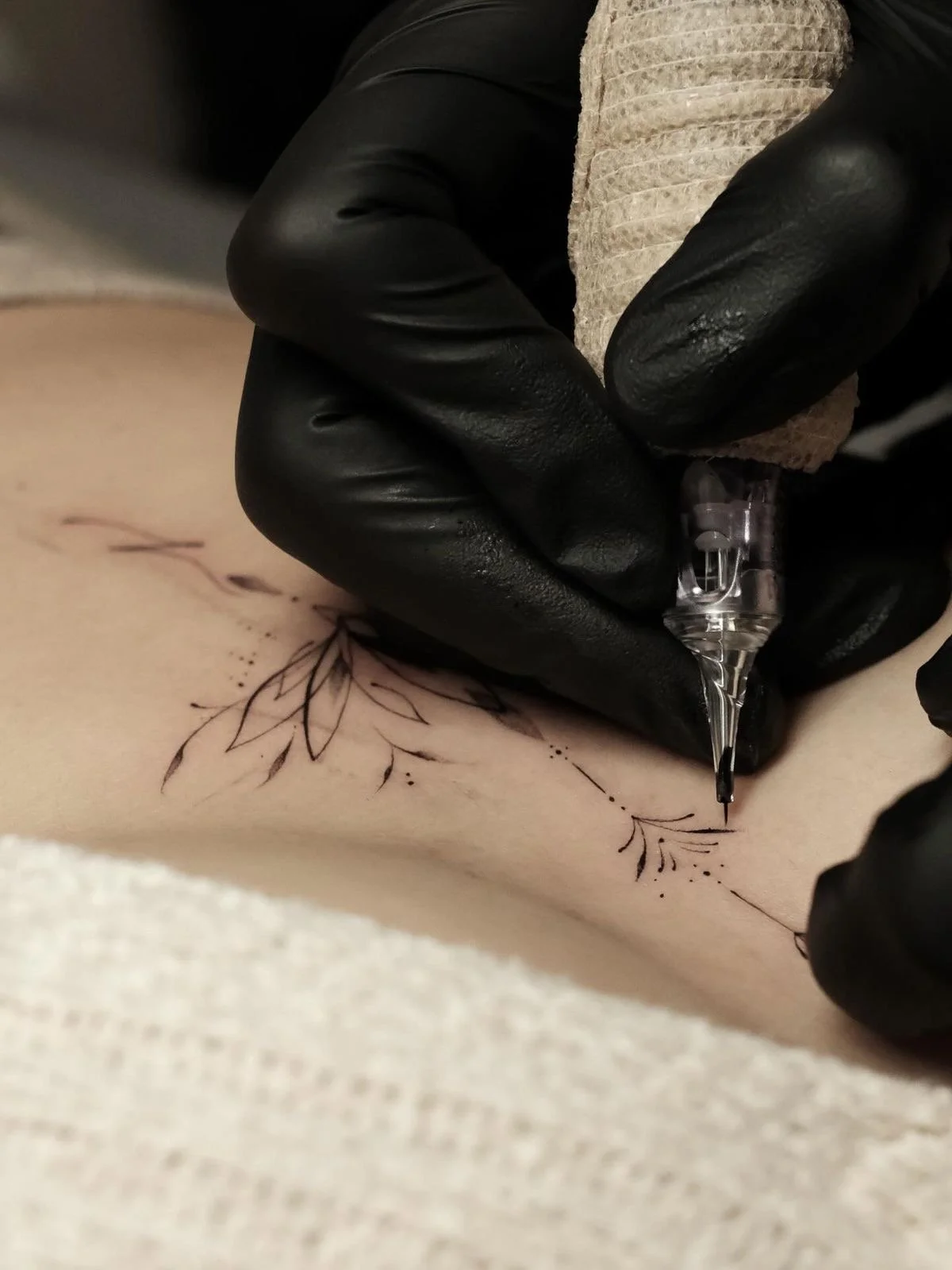 A person wearing black gloves is tattooing a delicate, black ink floral design on someone's skin.