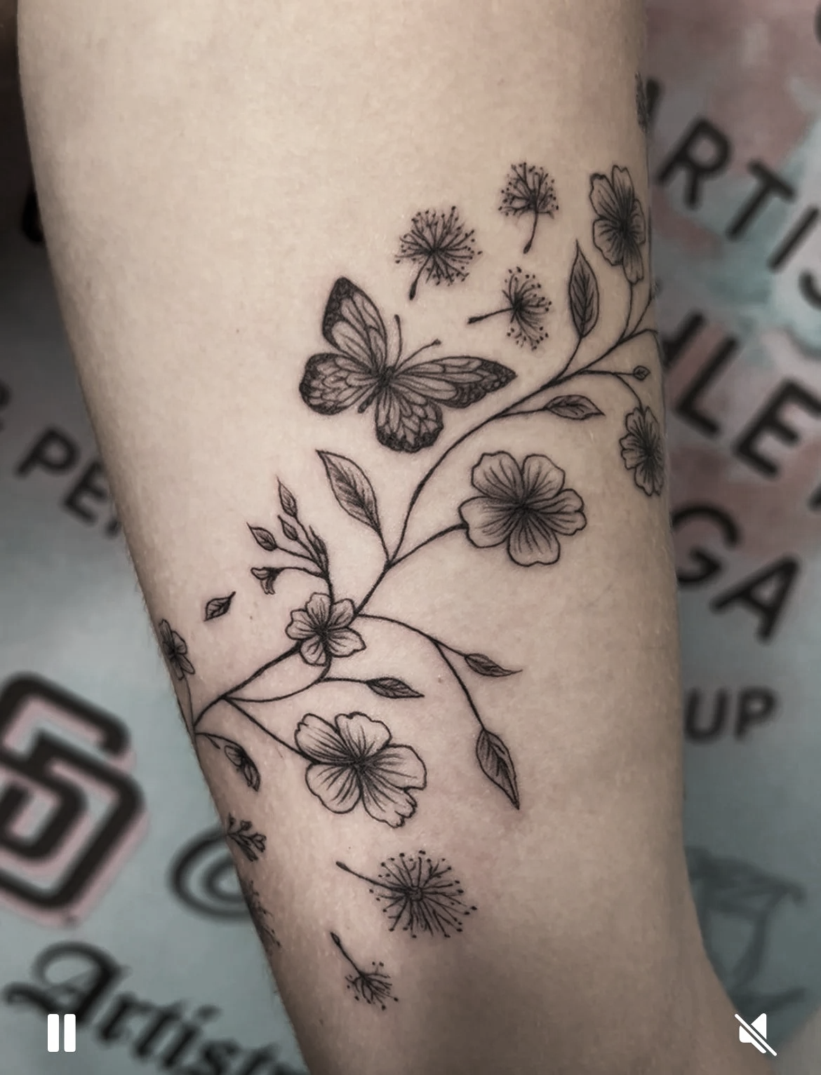 Black outline tattoo of a butterfly, dandelions, and flowers on forearm.