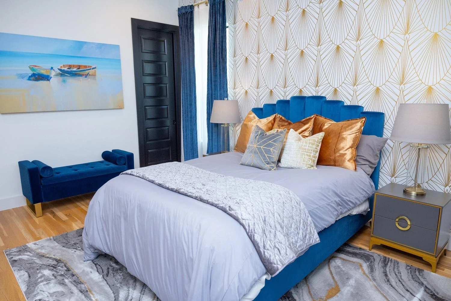 Modern bedroom with a blue upholstered bed and matching bench, gold and orange accent pillows, dark door, artwork of boats on water, striped and geometric wallpaper, wooden floors, and side lamps with gray shades.