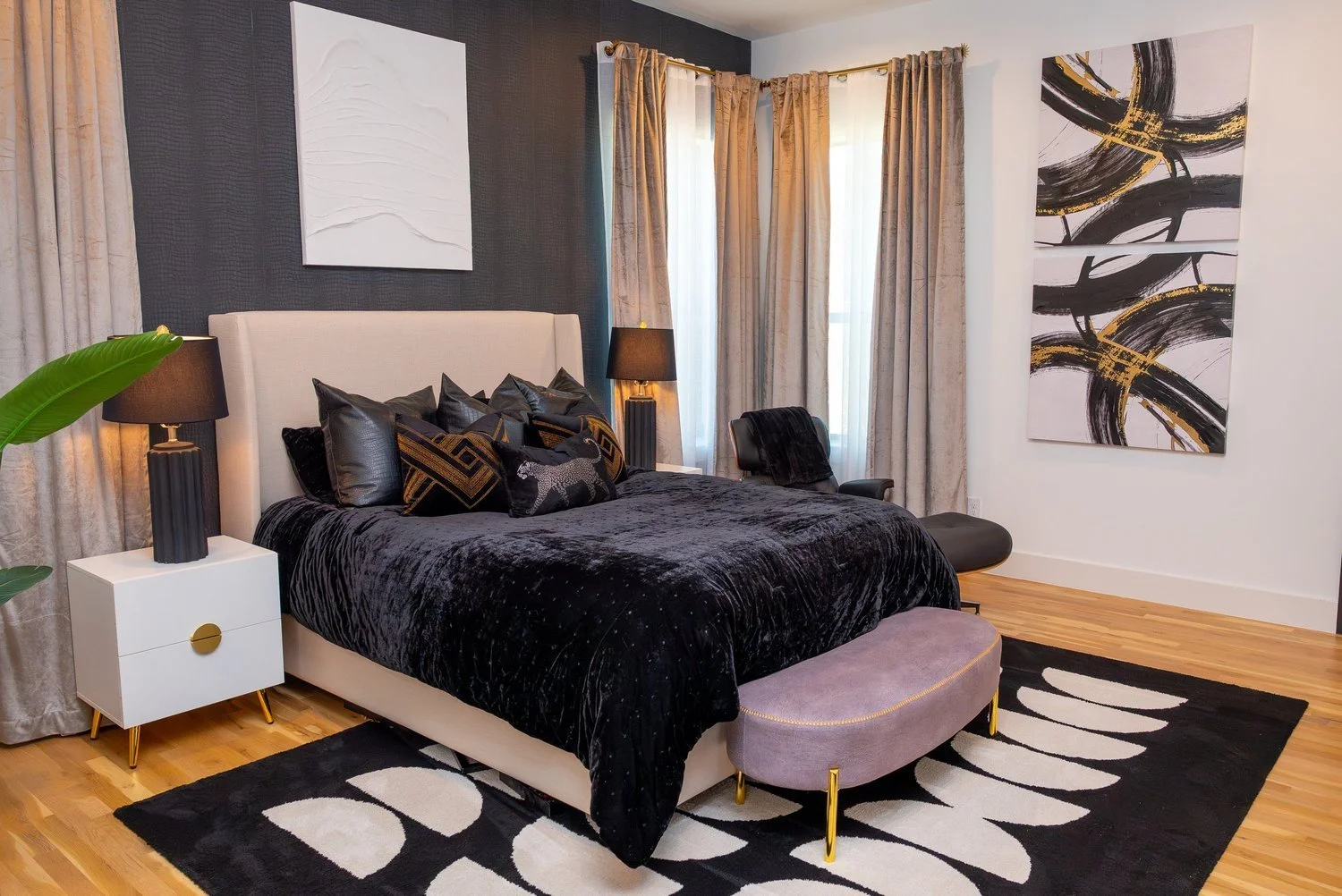 A modern bedroom with a black velvet bed, multiple black and patterned pillows, an off-white headboard, black side lamps, art pieces on the walls, curtains, a black and white area rug, a grey upholstered bench at the foot of the bed, and a black chair.