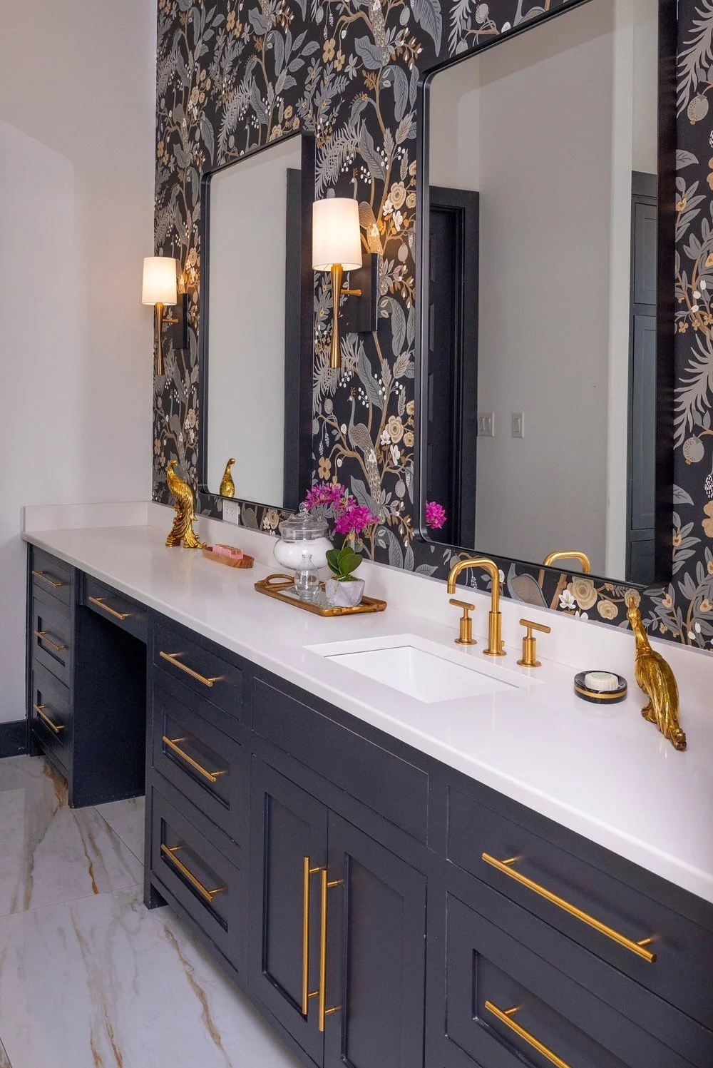 Elegant bathroom vanity with a dark blue cabinet, gold handles, white countertop, gold fixtures, large mirrors, floral wallpaper, and decorative gold peacocks and pink flowers.