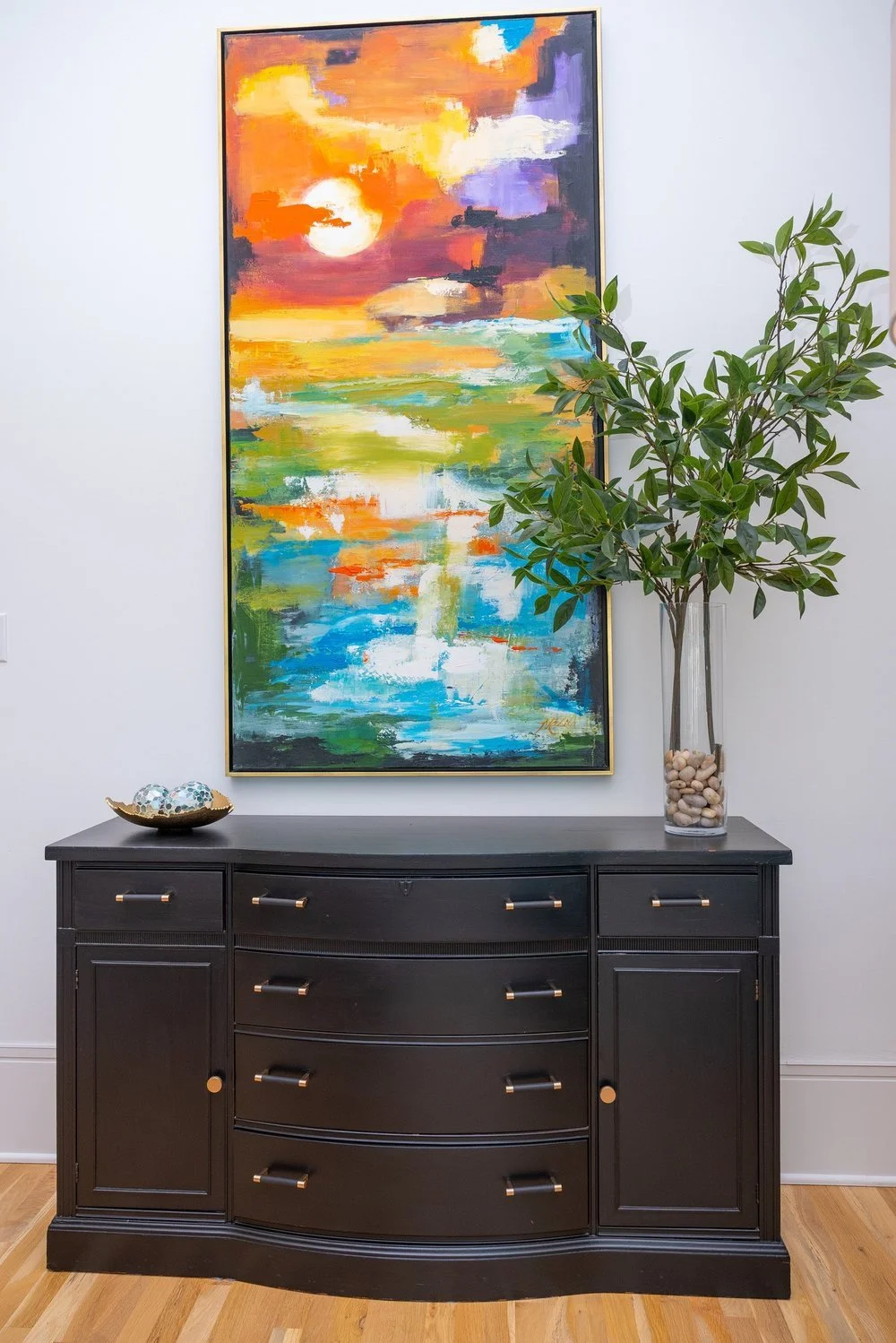 An abstract colorful painting with a sunset scene hangs above a black sideboard with drawers and cabinet doors. There is a tall glass vase with greenery and stones on the right side of the sideboard. A small decorative bowl with spherical objects is on the left side of the sideboard.