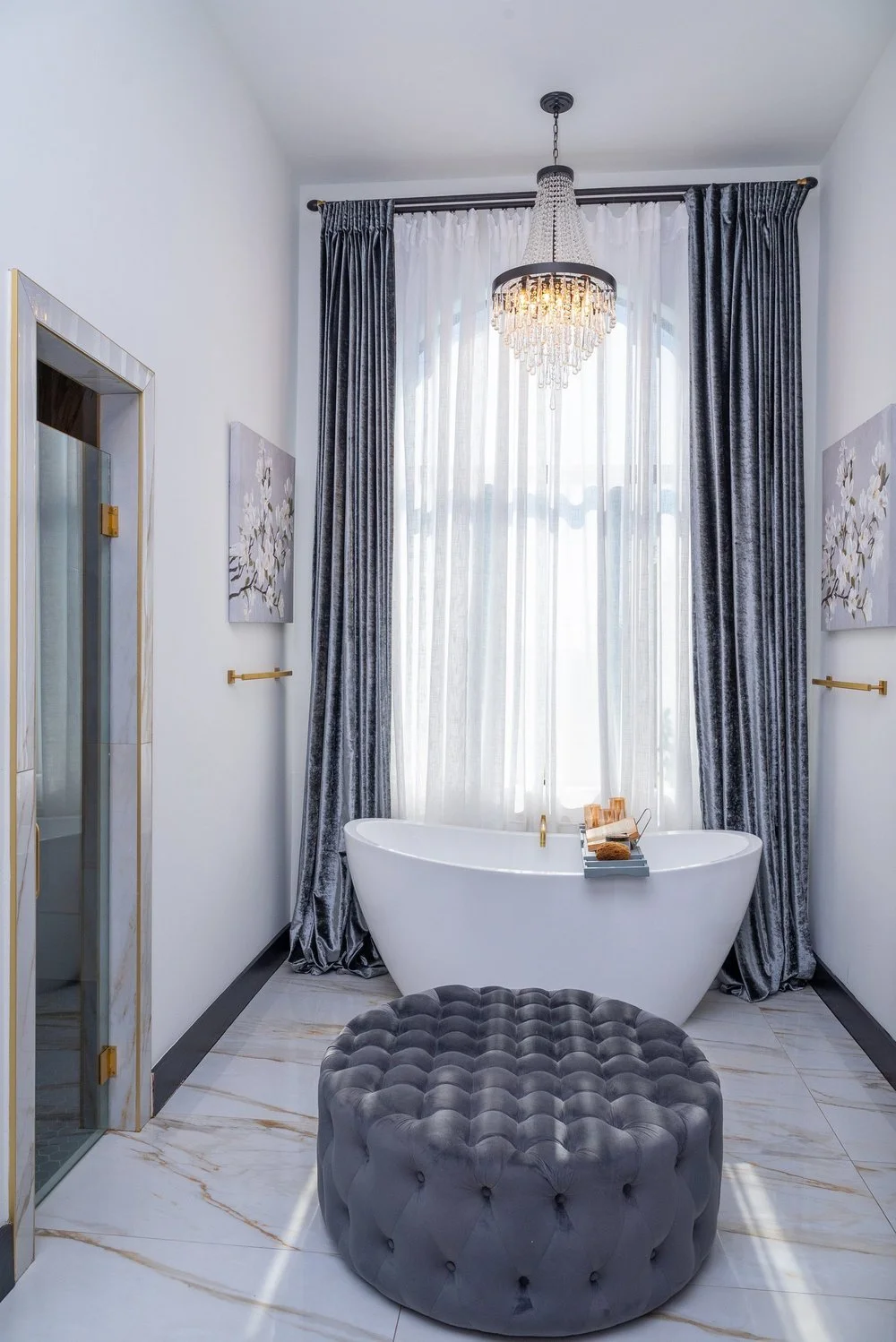 A luxurious bathroom with a freestanding bathtub in front of large window with sheer curtains and dark velvet drapes, a chandelier hanging from the ceiling, and a plush gray tufted ottoman on marble flooring.