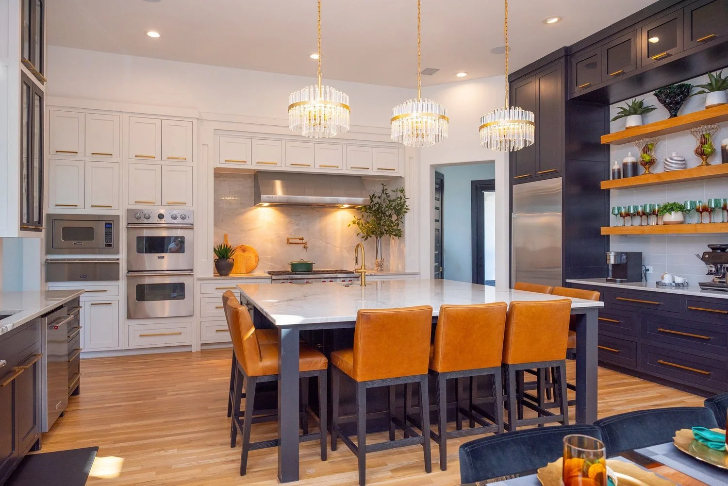 Modern kitchen with white upper cabinets, dark lower cabinets, a large marble island with four tan leather barstools, warm wood floors, and three crystal chandeliers hanging above the island.