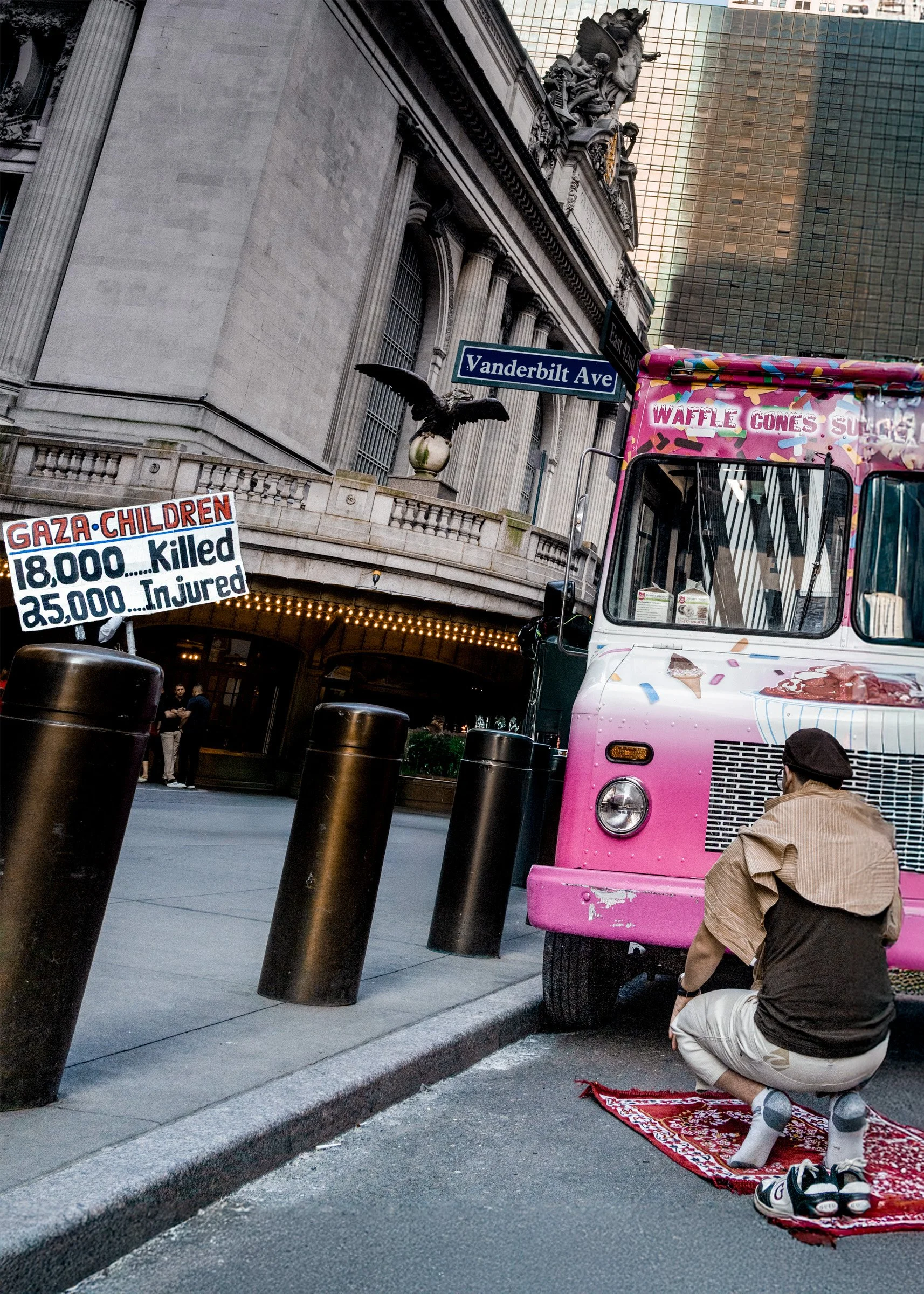 A man kneeling on a red rug facing a pink ice cream truck on a city street, with a sign referencing the Gaza children's death toll, and a Vanderbilt Ave street sign visible in the background.