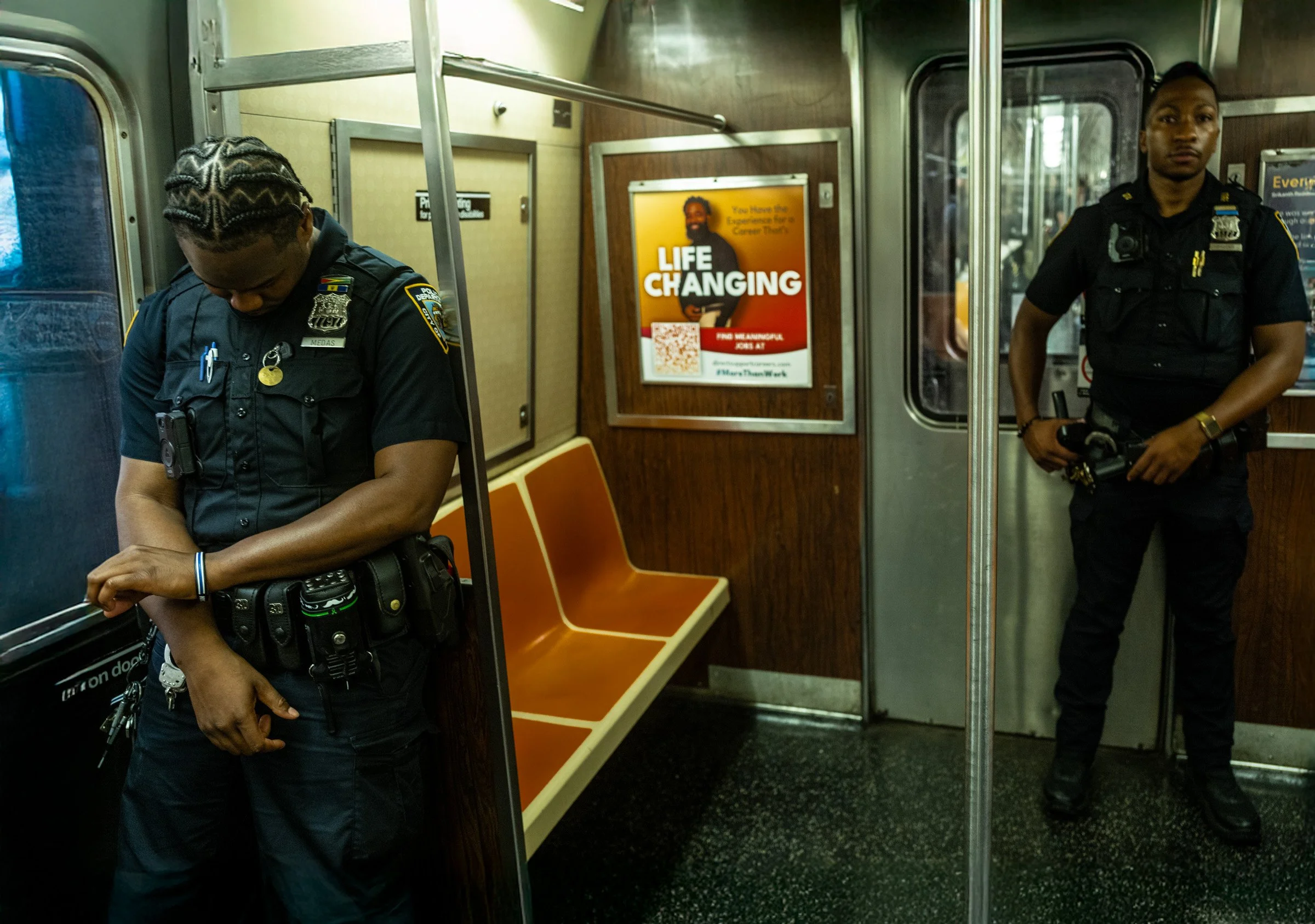 Two police officers inside a subway train. One officer is looking down at the ground, while the other is standing near the door, looking directly at the camera. The train has orange seats, and there is a poster on the wall that reads 'Life Changing' 