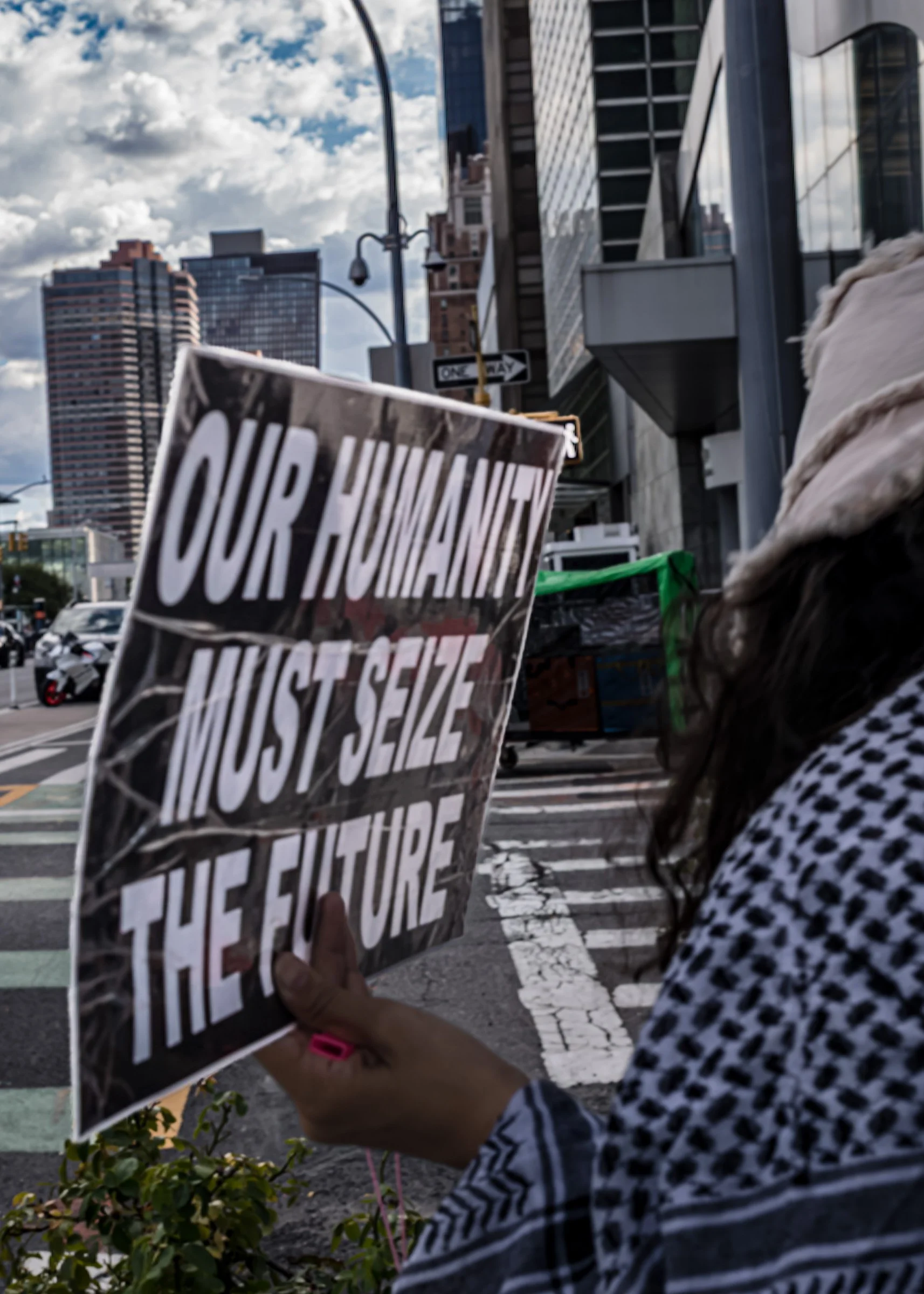 Person holding a sign that reads 'OUR HUMANITY MUST SEIZE THE FUTURE' on a city street with tall buildings and a partly cloudy sky.