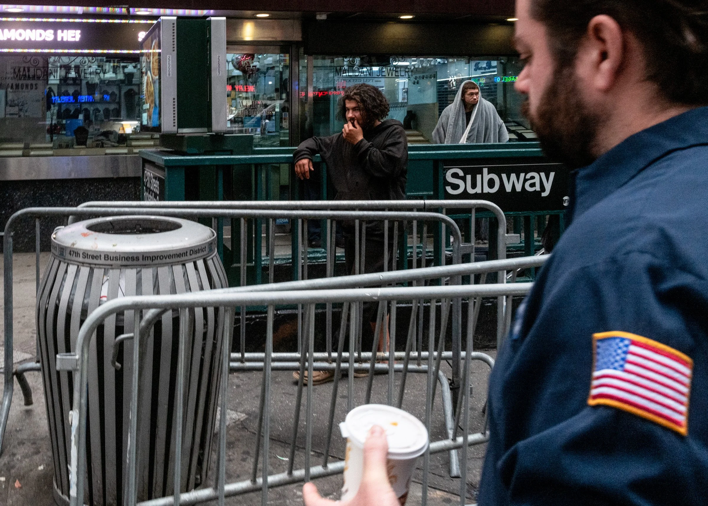 A man in a dark jacket leaning on a subway entrance railing, with two women in the background; one in a gray hoodie with a blanket over her head and the other in black, a police officer in uniform on the right holding a cup, placed near a metal trash