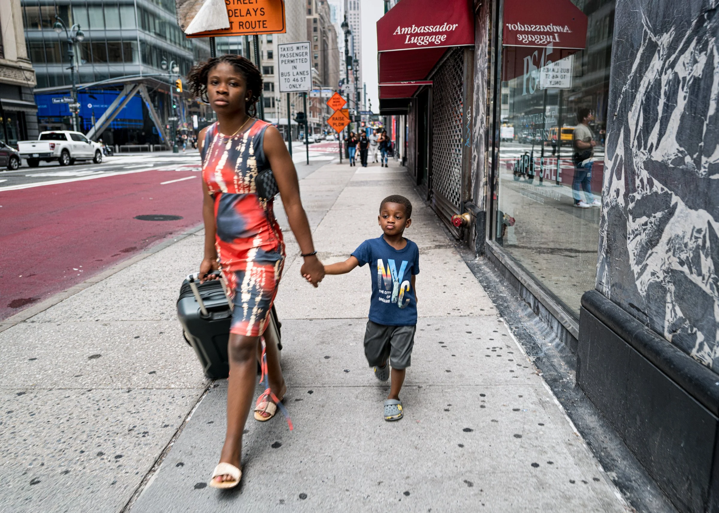 A woman and a young boy walking hand in hand on a city sidewalk. The woman is wearing a colorful, patterned dress and sandals, while the boy is dressed in a blue t-shirt and shorts. The city street has cars, traffic signs, and storefronts, with pedes