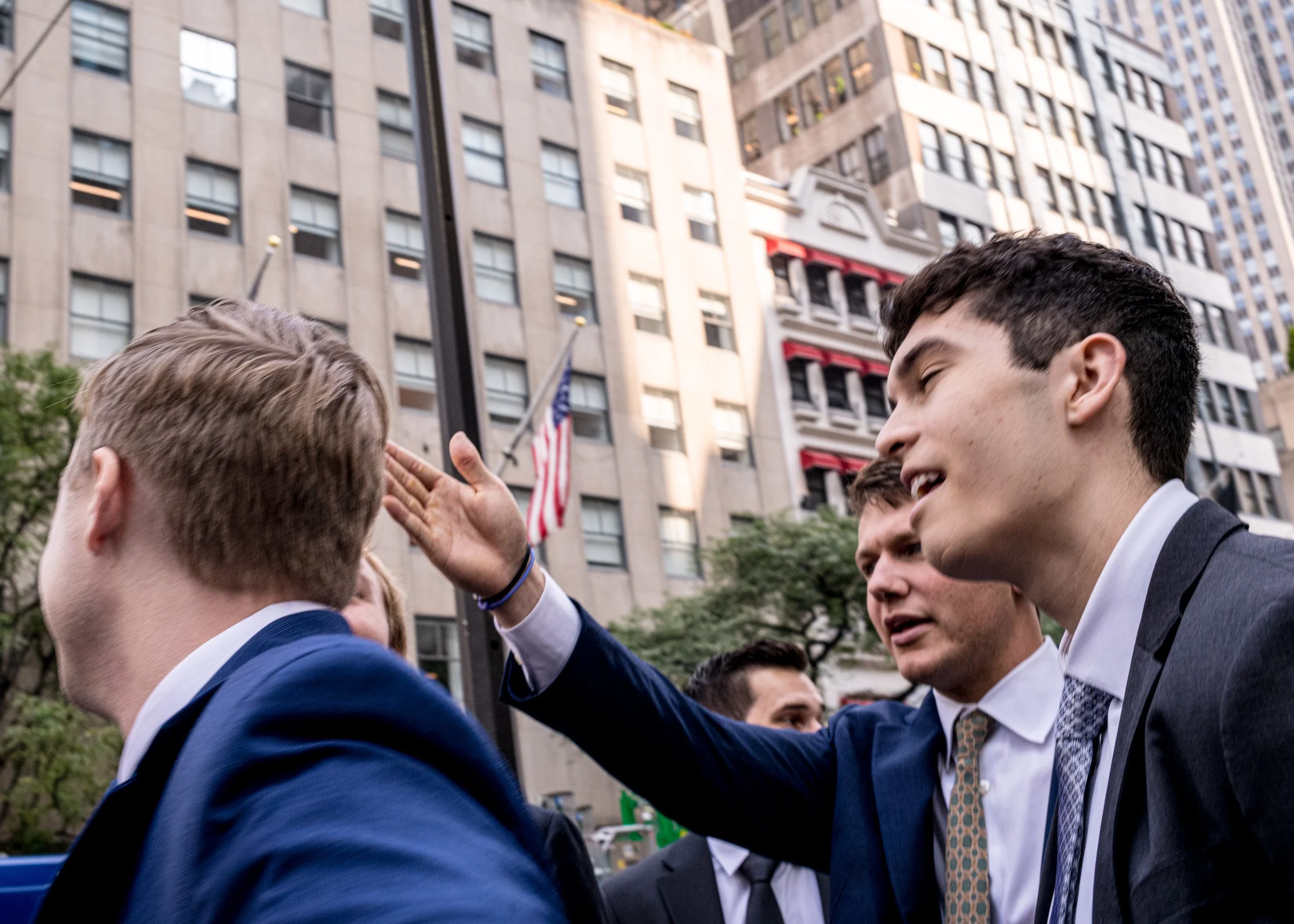 A group of men in suits having a discussion outdoors in front of tall buildings, with one man raising his hand as if greeting or making a gesture, and an American flag in the background.