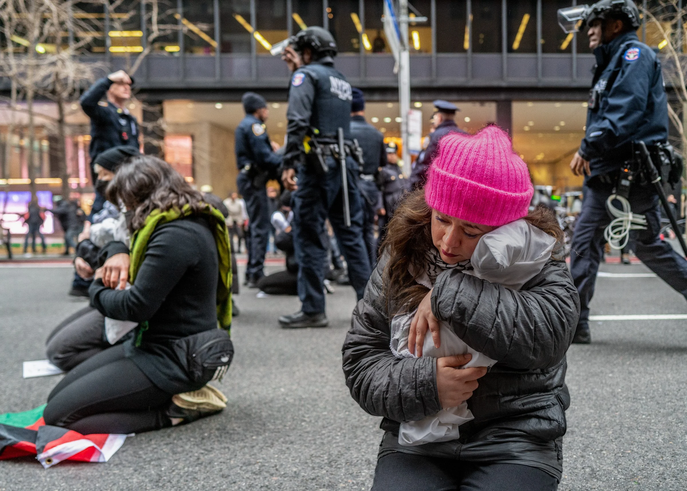 Two women kneel on the street, hugging themselves with distressed expressions during a protest. Police officers stand in the background, some saluting, amidst a city setting.