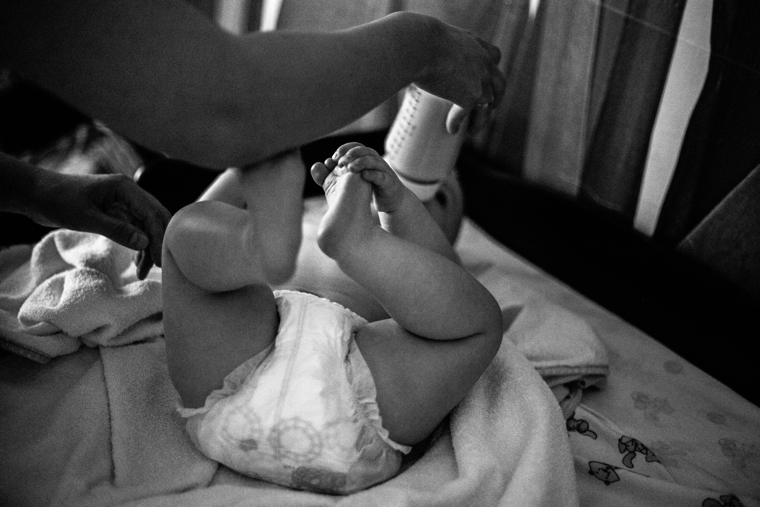 A baby lying on a bed, grabbing their feet with hands, with an adult's hand near them, in a black and white photo.