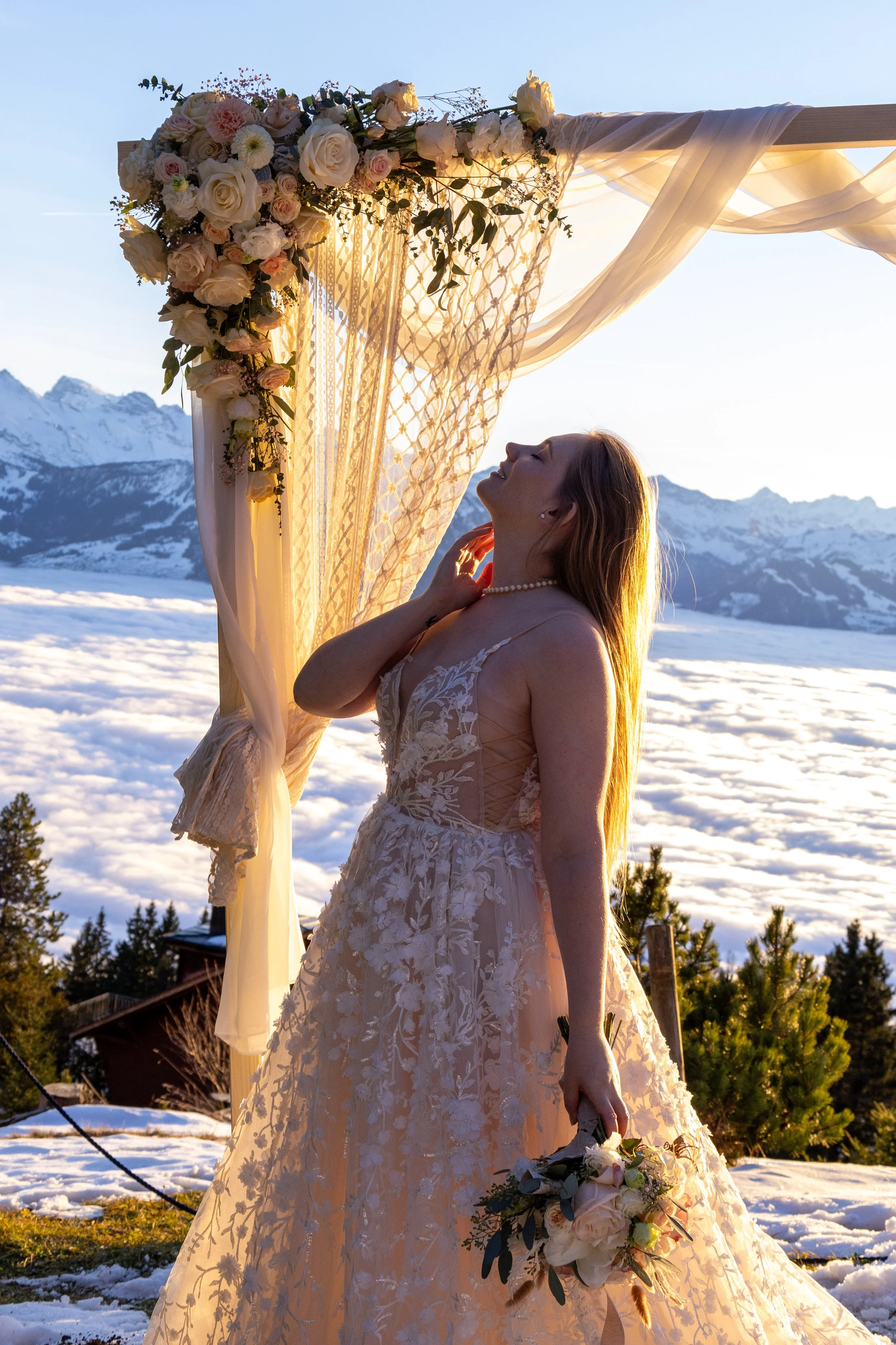 Braut in einem weißen Hochzeitskleid mit Blumenstrauß steht unter einem Blumenbogen vor Bergen und Wolken bei Sonnenuntergang.